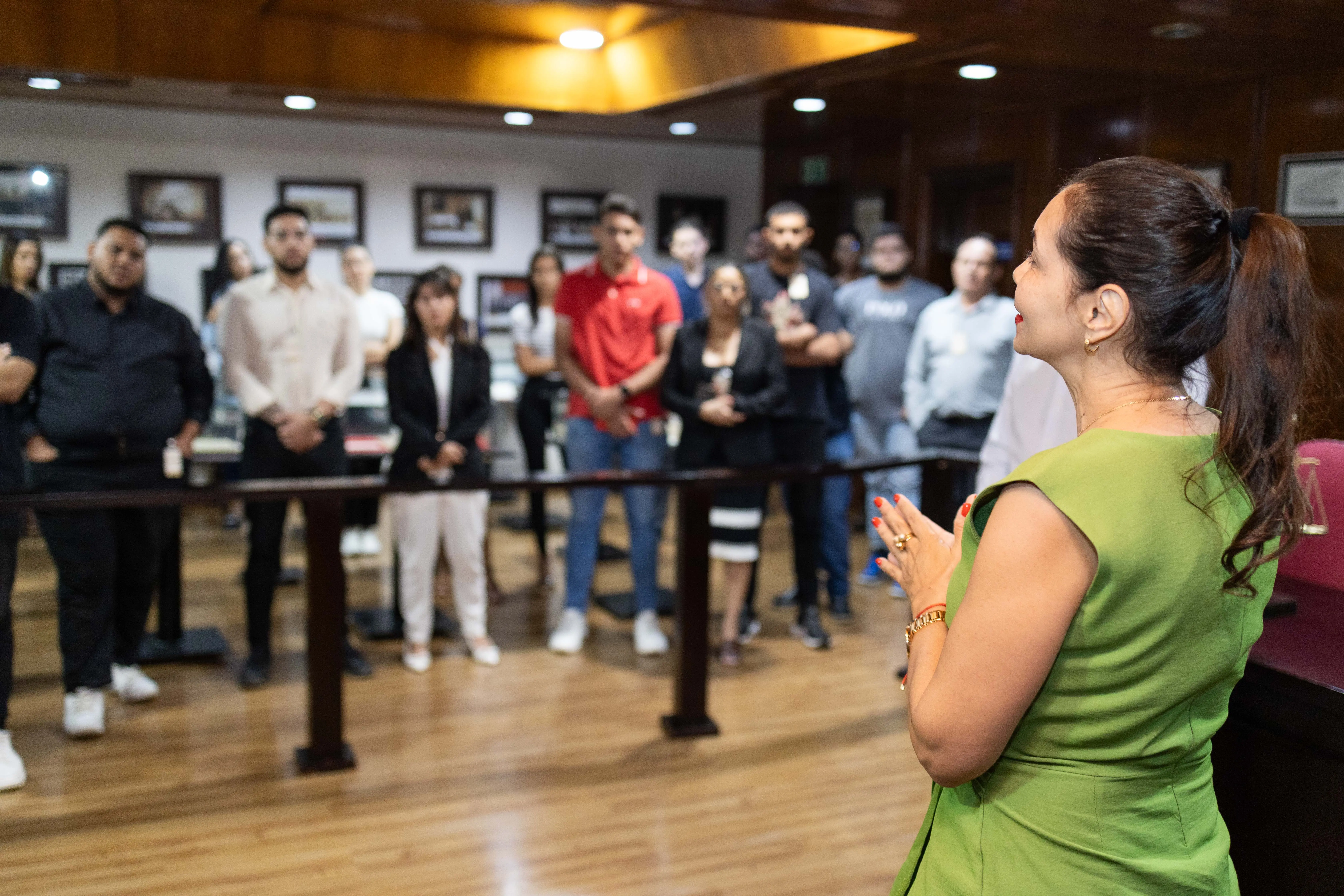 A juíza Anna Paula aparece de costas para a câmera enquanto fala com o grupo de 42  acadêmicos da Fasipe. Ela veste vestido verde folha e gesticula enquanto fala. Todos estão em pé. O ambiente é o Espaço Memória do PJMT, tem paredes de madeira escura e iluminação suave.