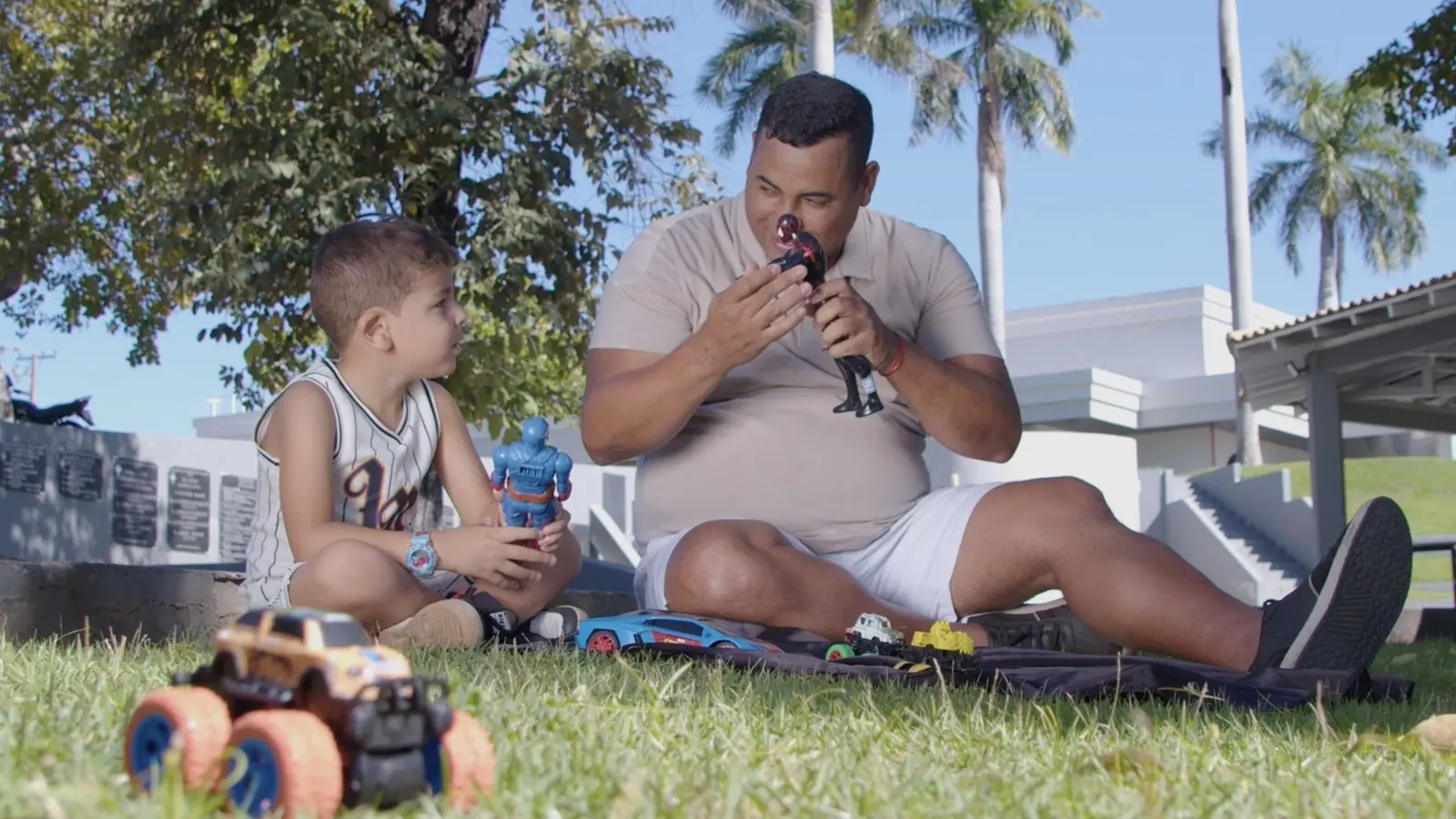 Pai e filho brincam sentados na grama em um dia ensolarado. O pai interage com um boneco, enquanto o menino observa atento segurando outro brinquedo. Ao redor, há carrinhos e um cenário com árvores e prédio ao fundo. 