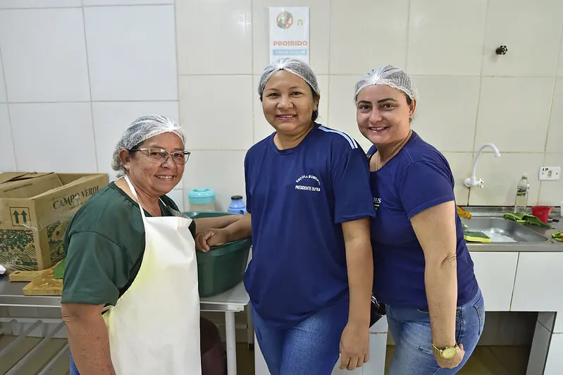 Quatro mulheres que trabalham na cozinha da Escola 11 de Agosto posam para a foto sorrindo. Elas usam touca e avental.