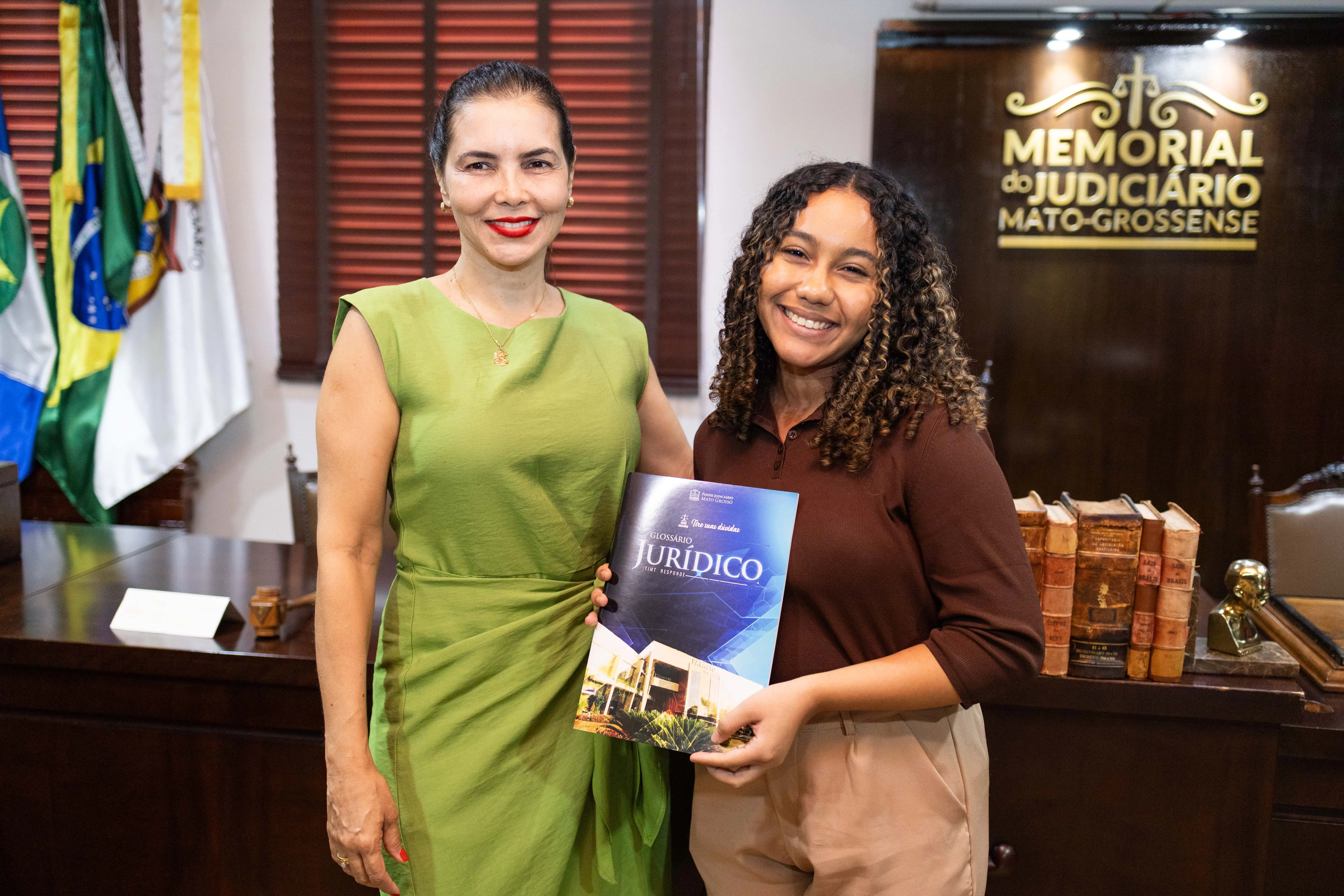A juíza Anna Paula está sorridente e posa ao lado da jovem estudante universitária, Anne Heloísa, que também sorri e segura um livreto azul intitulado "Glossário Jurídico". Ao fundo, à direita, há uma mesa de madeira escura com livros antigos e letreiro dourado com os dizeres "Memorial do Judiciário