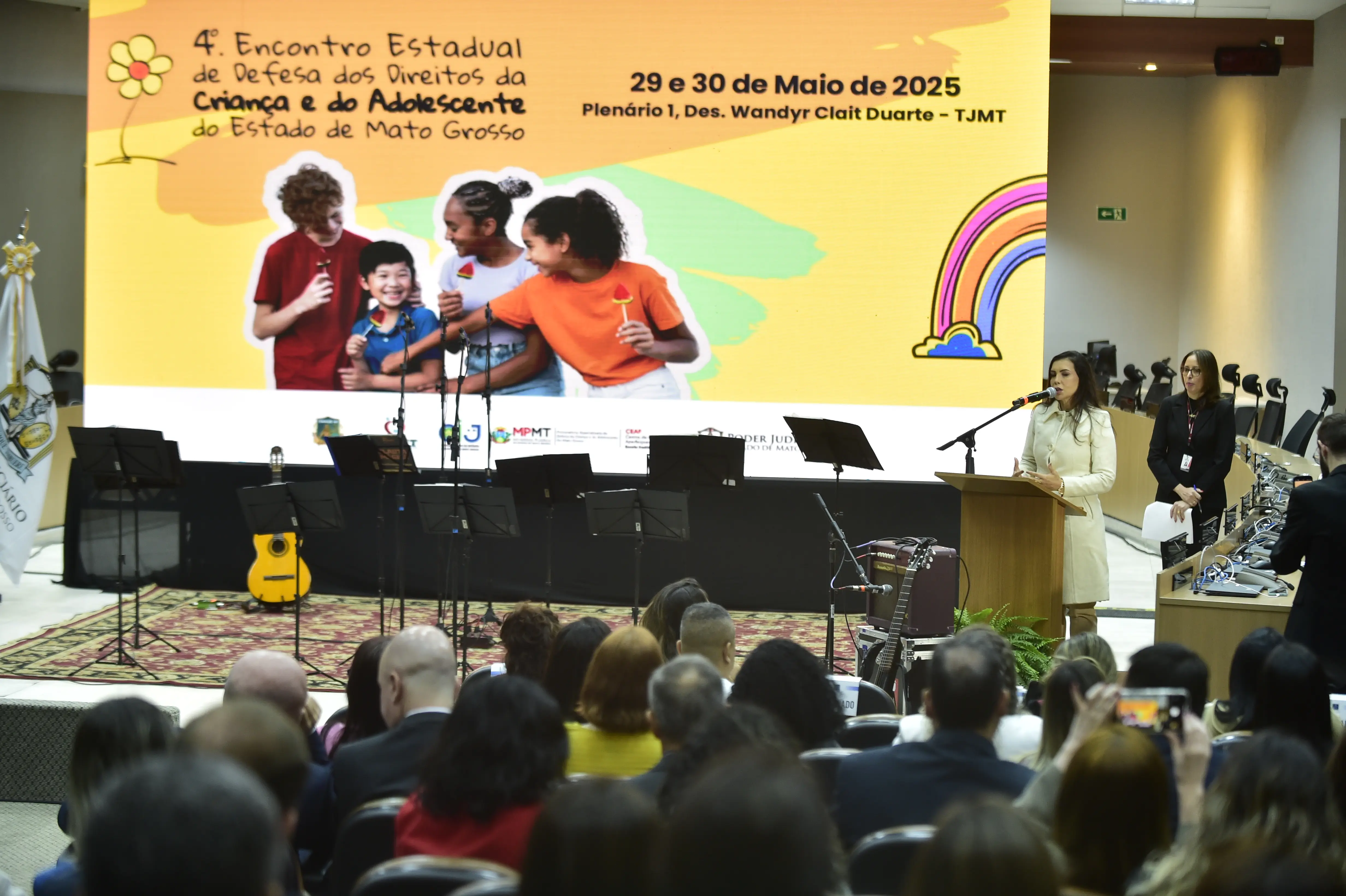 Mulher discursa em palco no 4º Encontro Estadual de Defesa dos Direitos da Criança e do Adolescente de MT. Ao fundo, telão colorido com imagem de crianças e informações do evento. Auditório cheio e instrumentos musicais ao lado.