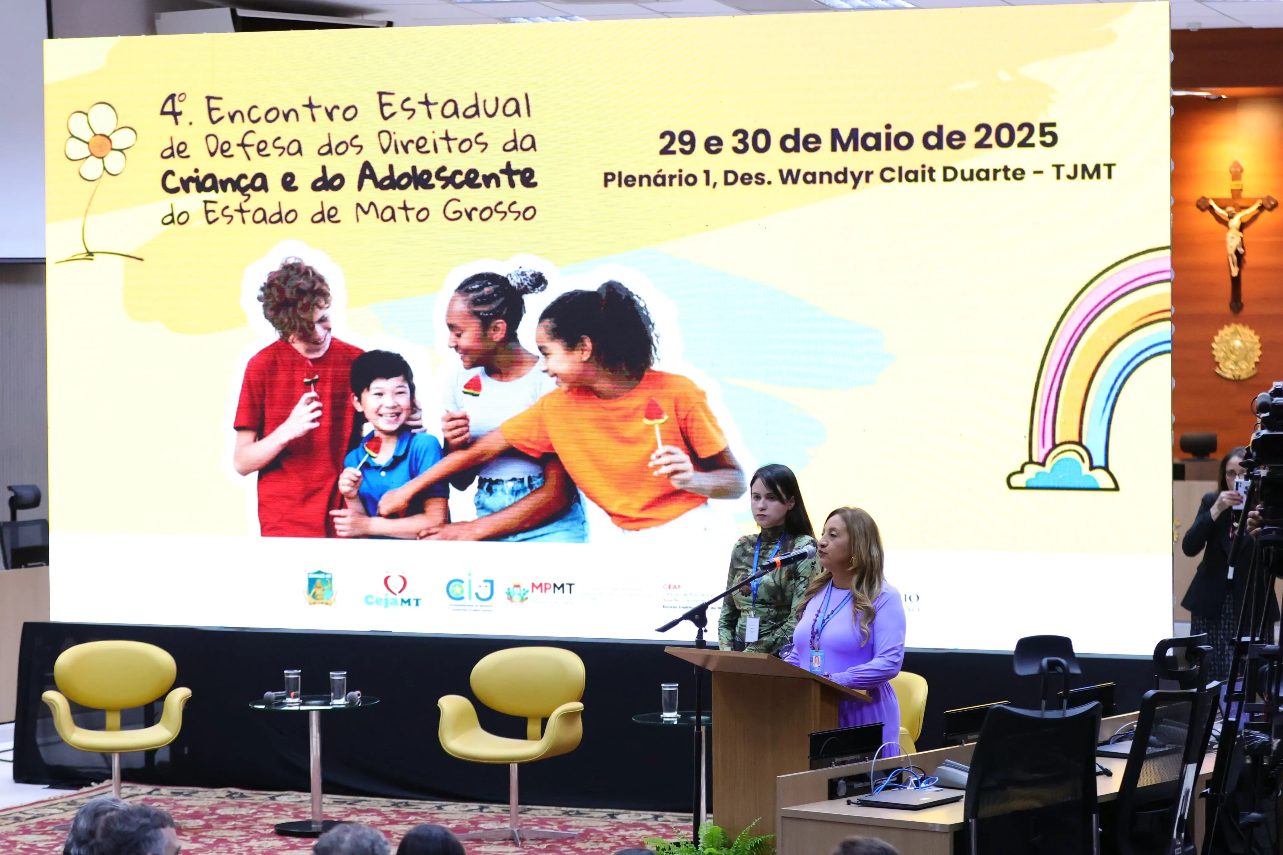No centro, uma mulher de cabelos loiros e roupa roxa está em um púlpito. Atrás dela, um grande telão exibe "4º Encontro Estadual de Defesa dos Direitos da Criança e do Adolescente do Estado de Mato Grosso" e "29 e 30 de Maio de 2025", com a imagem de crianças e um arco-íris. 