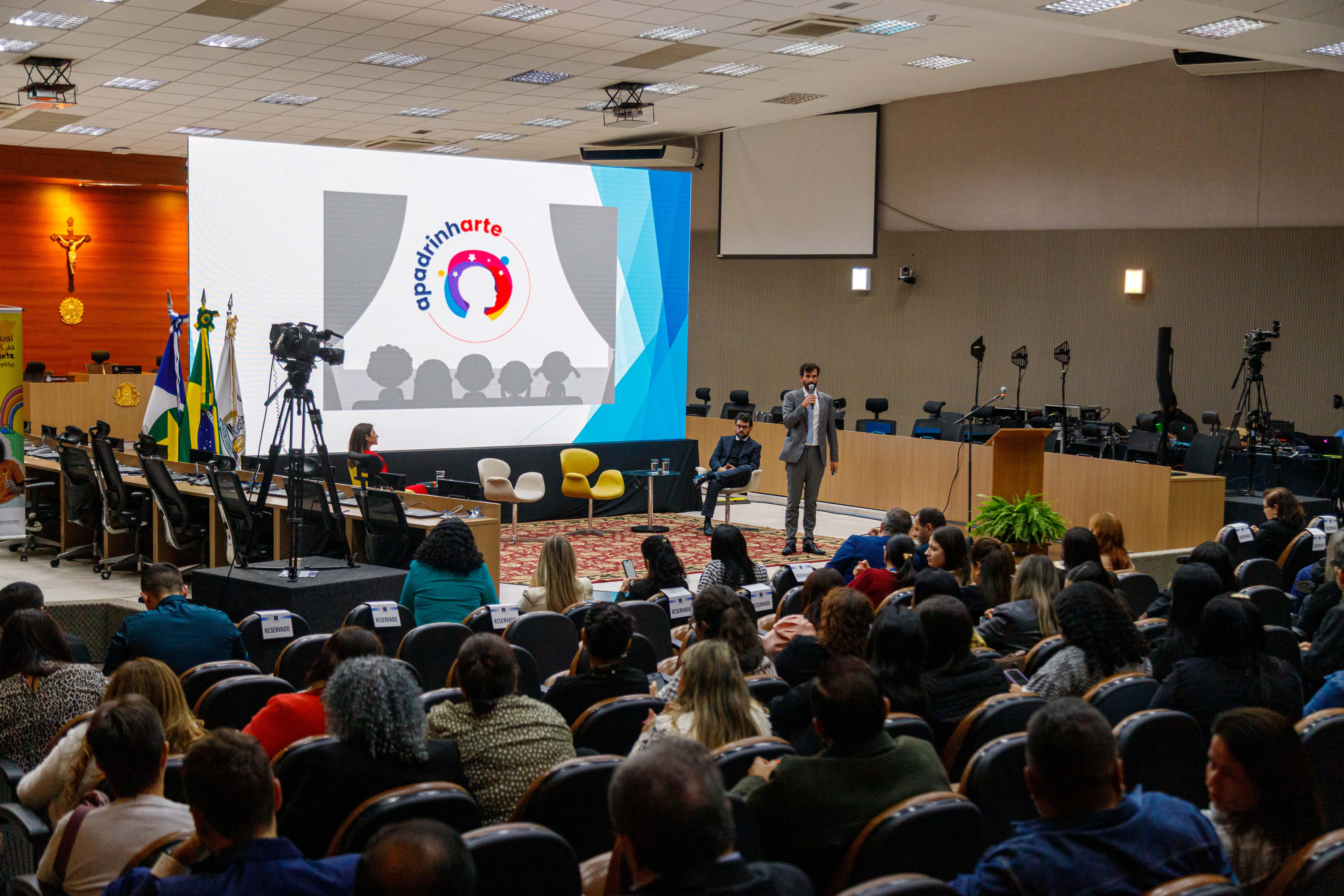 Auditório lotado com plateia atenta a uma apresentação. Palestrante em pé fala ao microfone diante de telão com texto e bandeiras do Brasil e Mato Grosso. Há câmeras filmando o evento e iluminação clara.