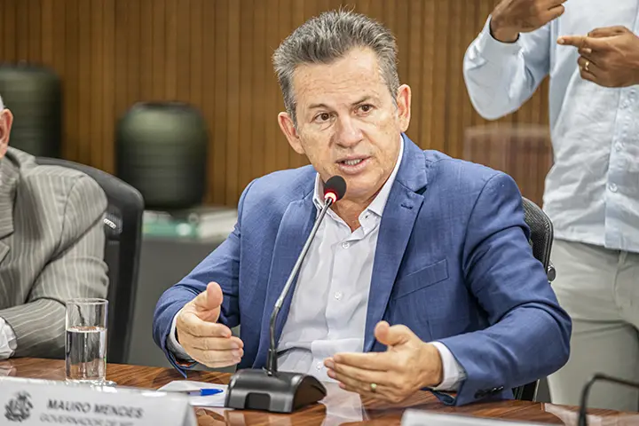 Foto em plano fechado do governador Mauro Mendes, vestindo um terno azul, gesticulando enquanto fala em um microfone sobre a mesa. Atrás dele, aparece um intérprete de Libras. O governador é um homem branco, de cabelos lisos e grisalhos. 