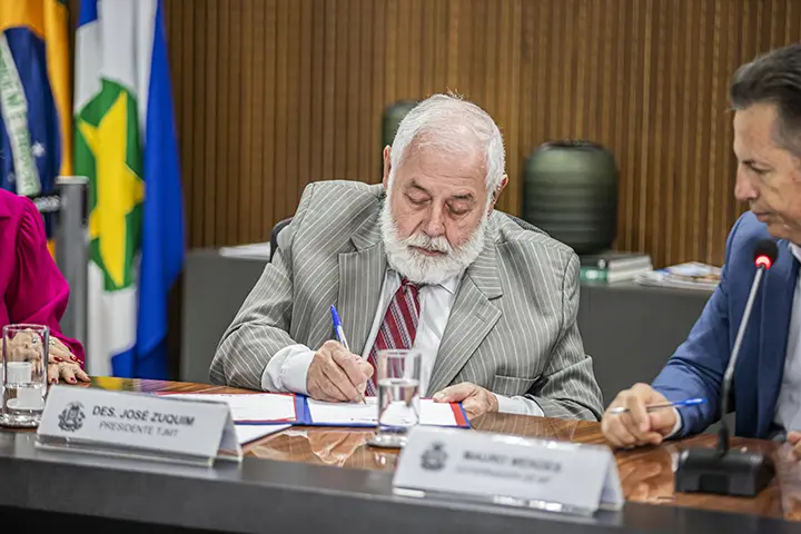 Desembargador José Zuquim assinando um documento na mesa de reunião do Palácio Paiaguás. O governador Mauro Mendes, sentado ao lado, observa o ato. José Zuquim é um senhor com cabelo e barba brancos, usando camisa cinza, terno cinza listrado e gravata vermelha listrada. 