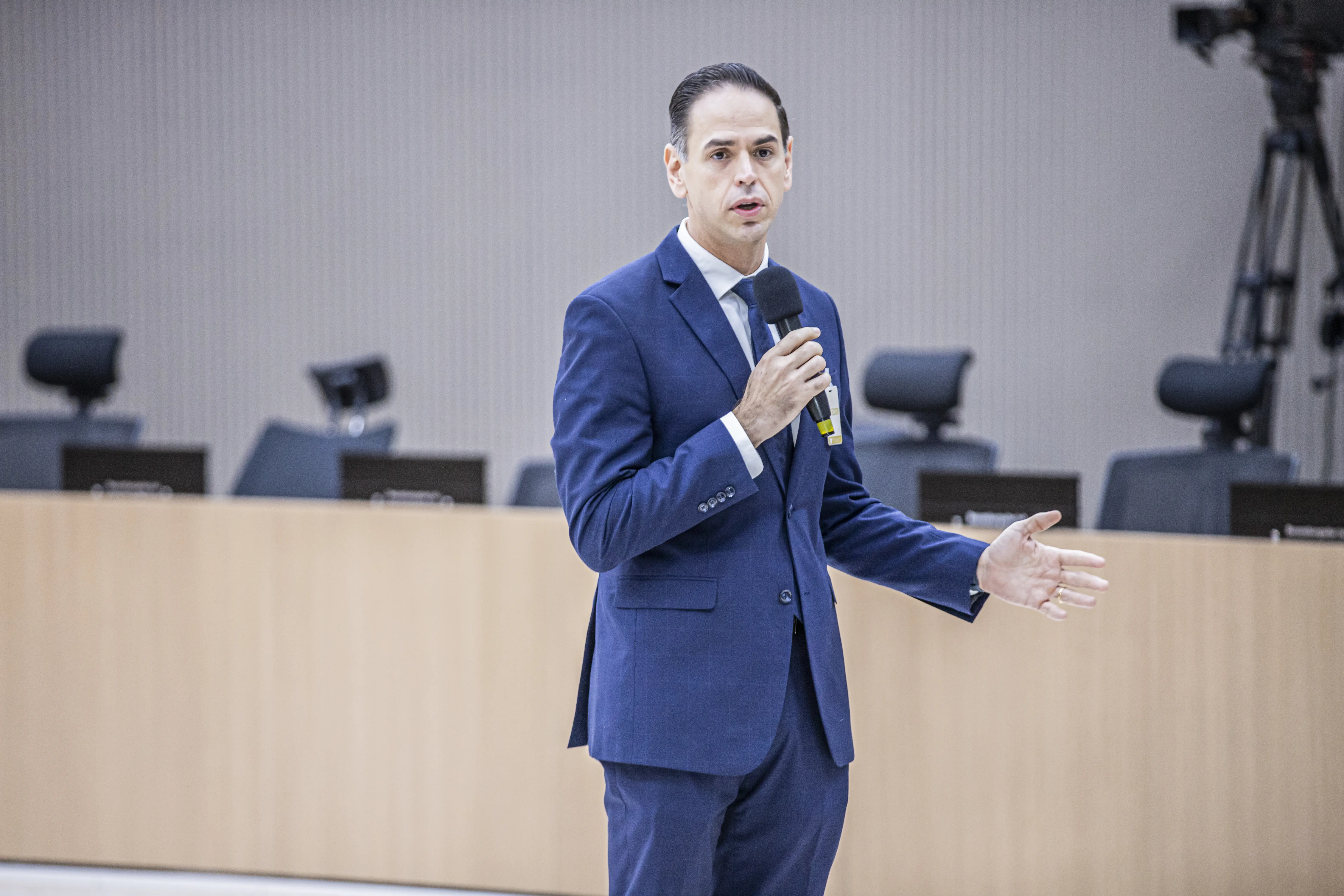 Homem de terno azul e gravata clara fala ao microfone com expressão séria e gesto de mão aberta. Está em um plenário com cadeiras giratórias e ambiente institucional ao fundo.