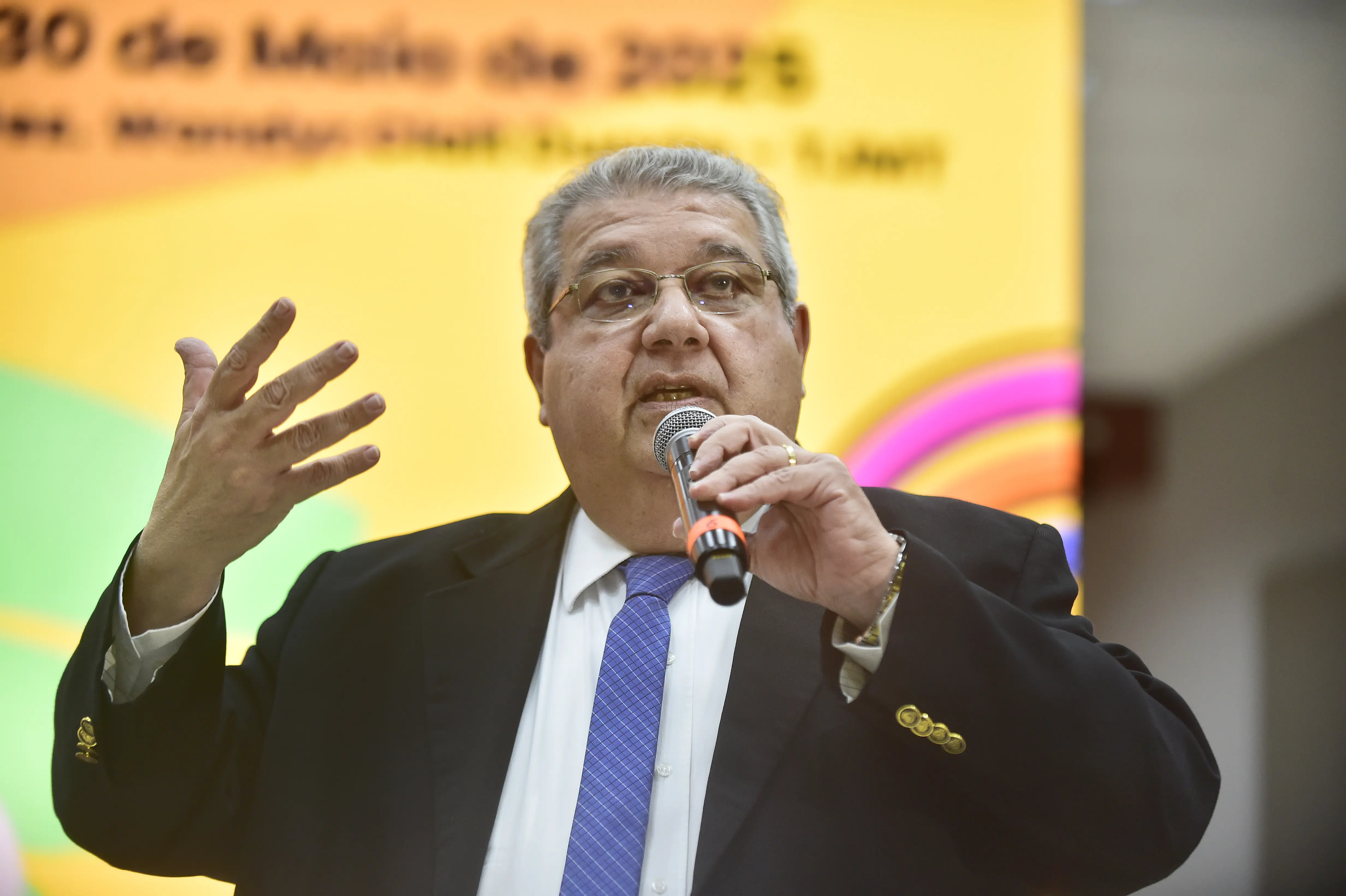 Homem grisalho, de óculos, terno escuro e gravata azul, fala em um microfone, gesticulando com a mão esquerda. Fundo amarelo desfocado com texto.