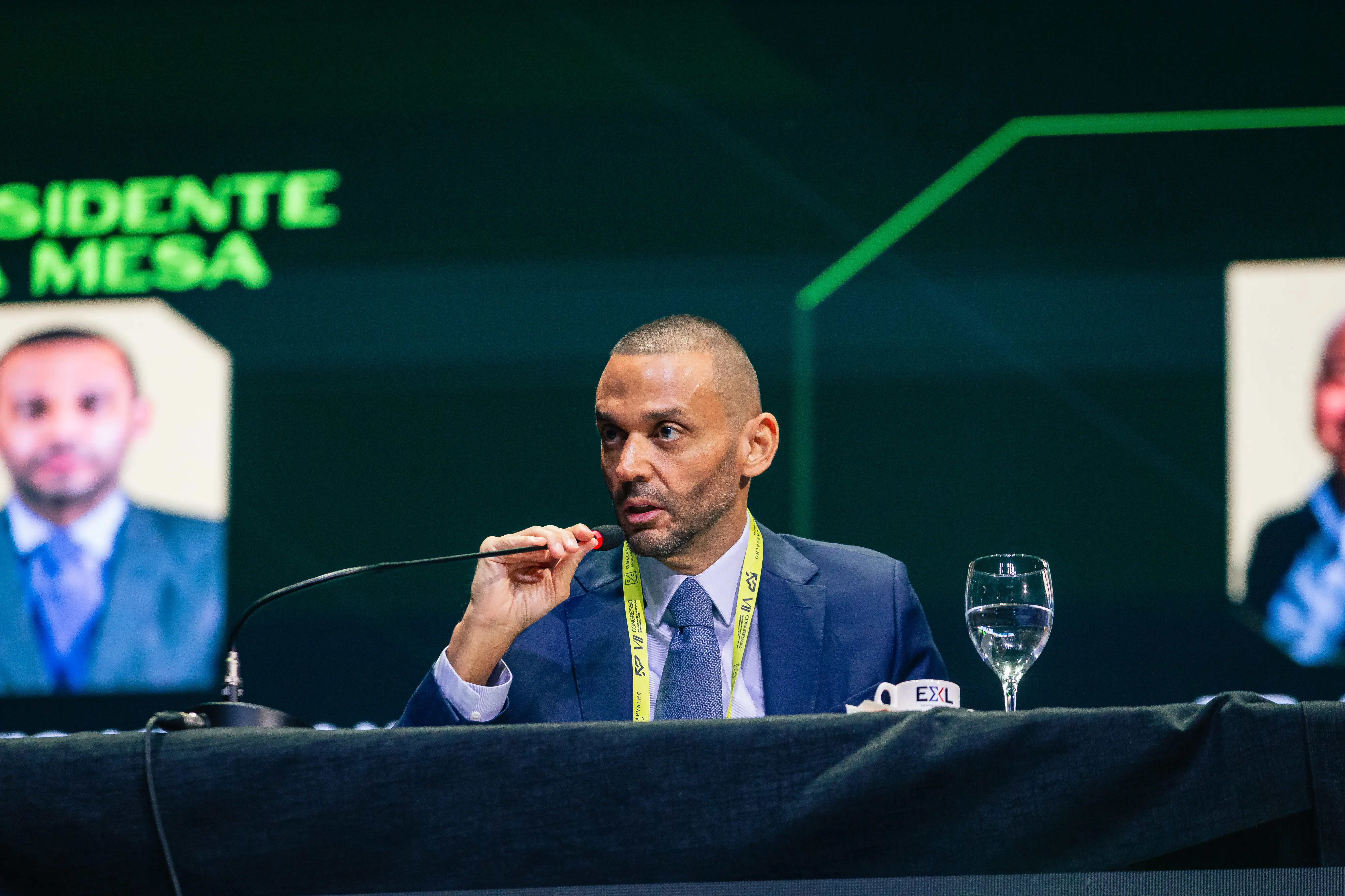 Juiz Flávio Miraglia durante participação no 7º Congresso de Reestruturação e Recuperação Empresarial. Ele está sentado e falando ao microfone. Ele é um homem pardo, magro, alto, com cabelos raspados, usando camisa branca, terno e gravata azul. 