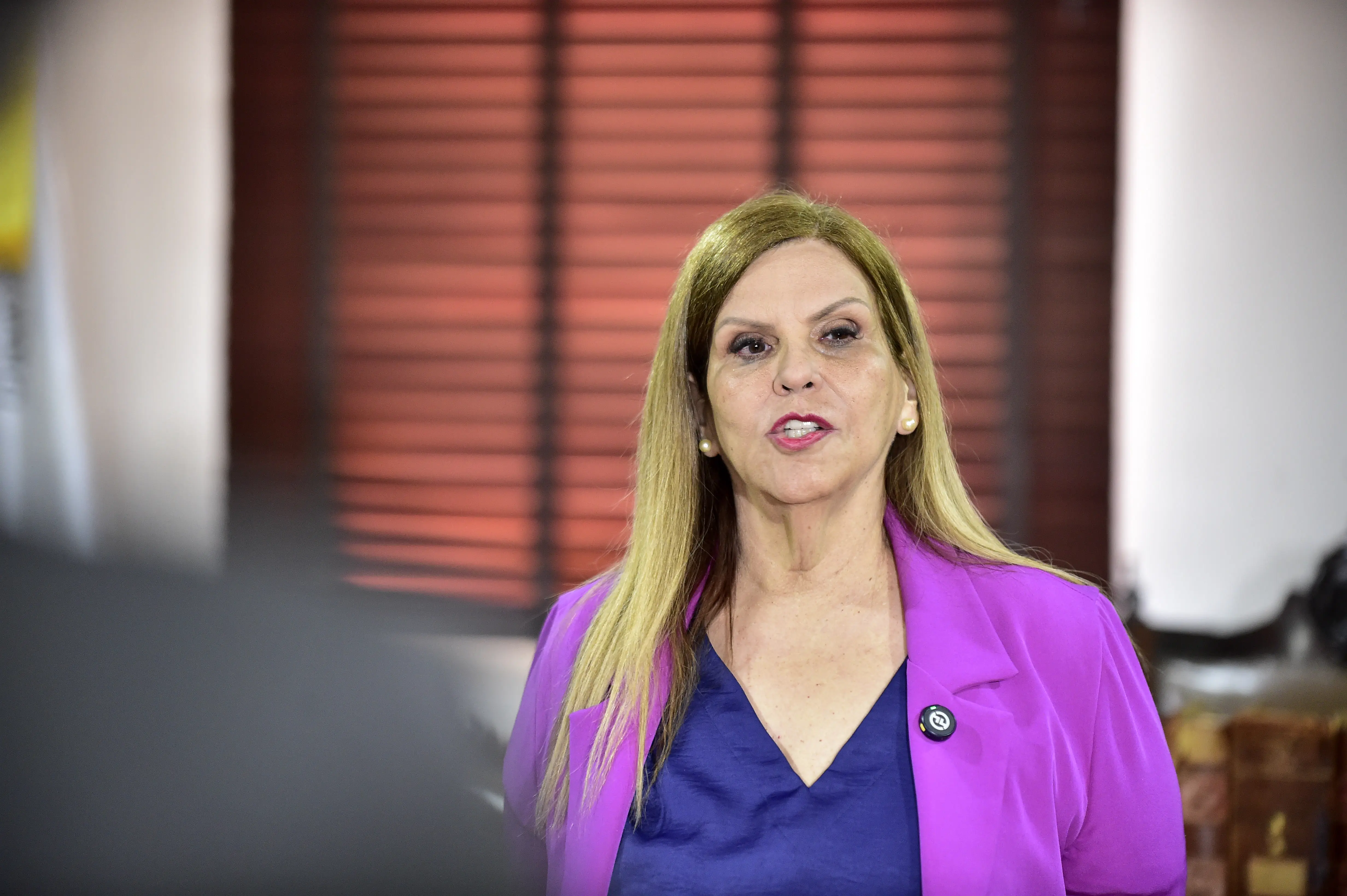 Mulher loira veste blazer roxo e blusa azul. Fala de forma expressiva, em destaque no ambiente iluminado, com janelas de madeira ao fundo. Sua postura transmite firmeza e segurança.