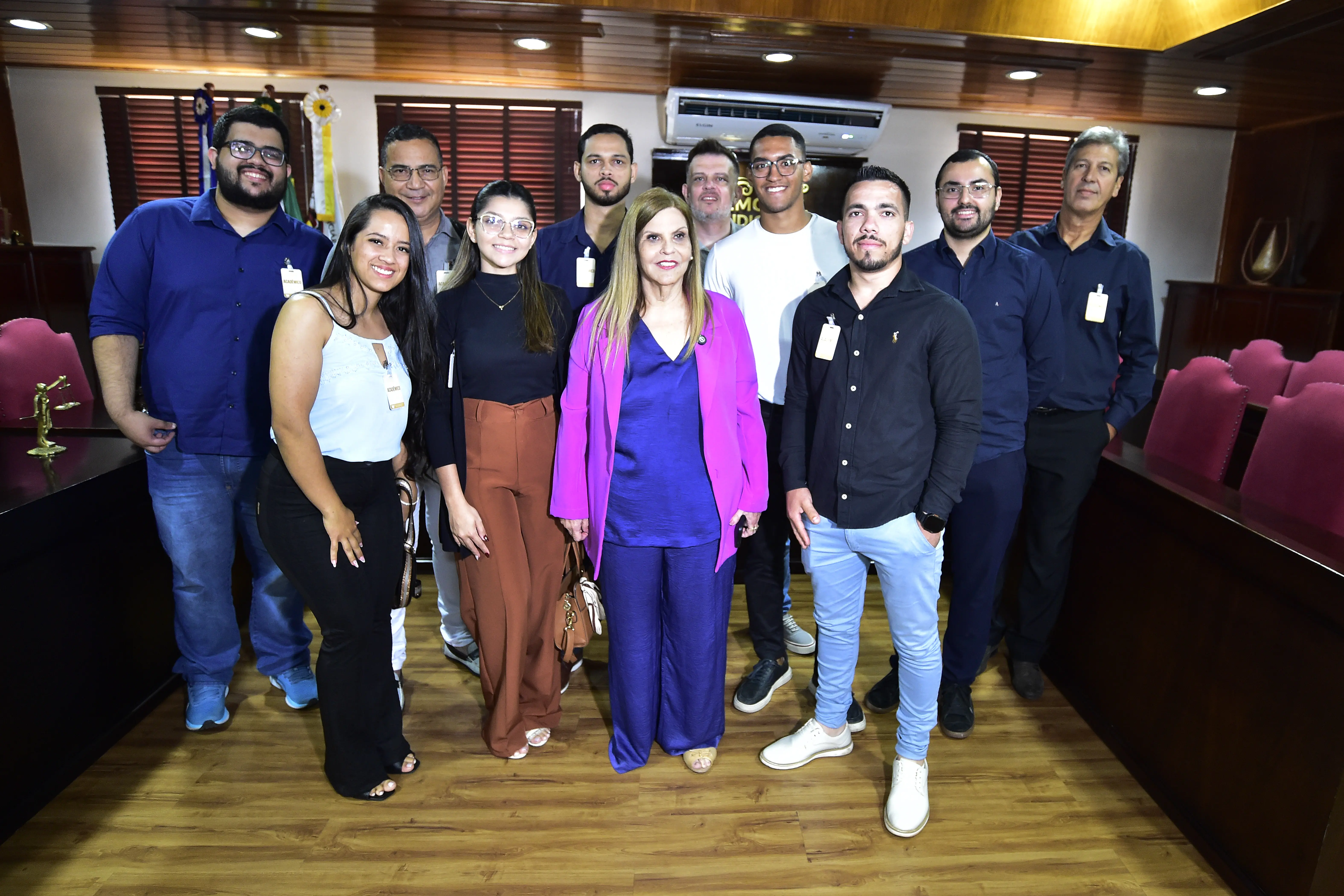 Grupo de estudantes posa ao lado de uma magistrada vestida de roxo no Memorial do Judiciário. Todos sorriem em clima de descontração, no espaço com móveis de madeira e cadeiras vermelhas.