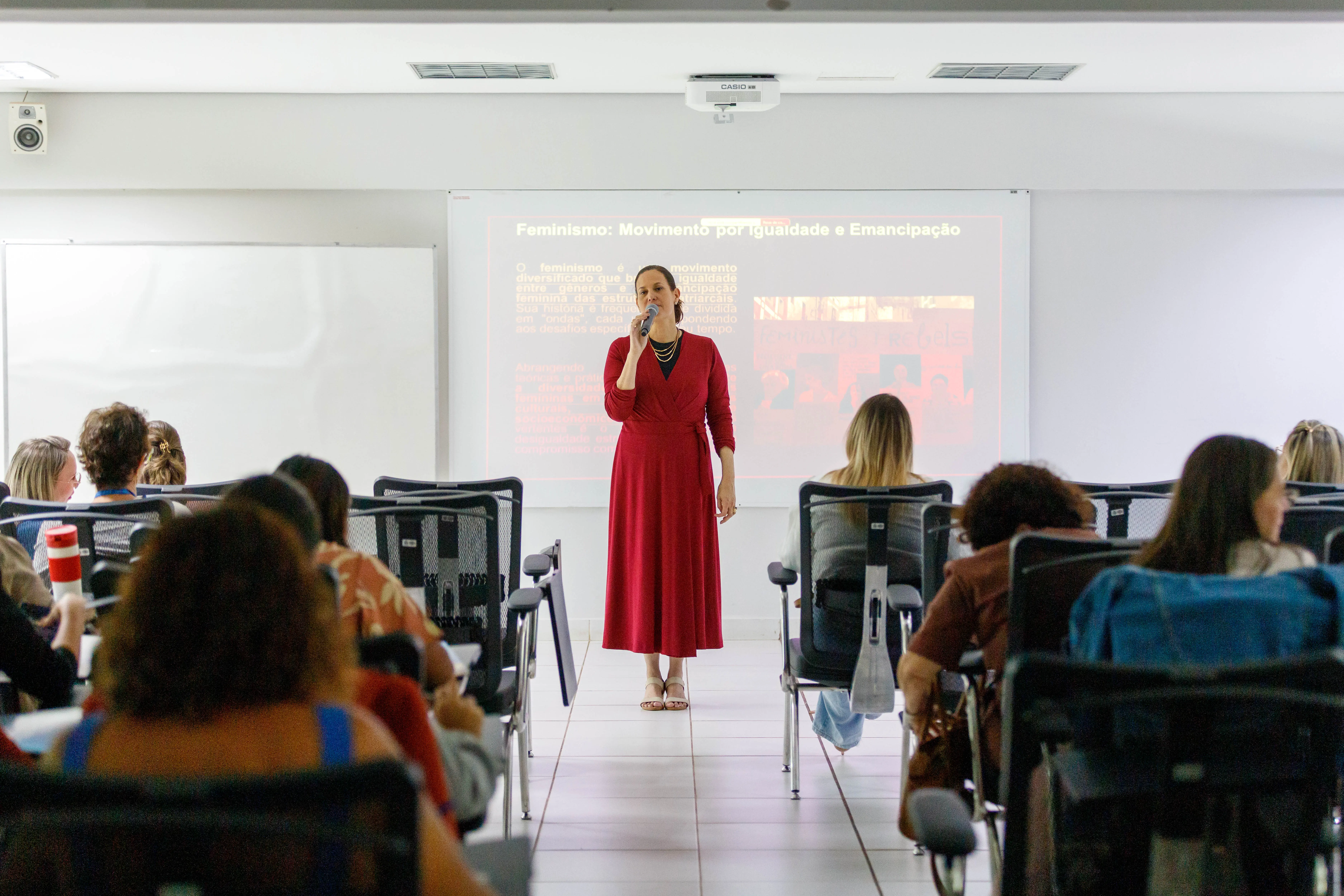 Palestrante de vestido vermelho fala ao microfone diante de telão com conteúdo sobre feminismo. Público está atento em sala com cadeiras pretas e iluminação clara