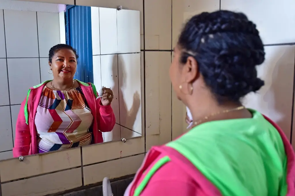 Mulher posa sorrindo para a foto, em frente a um espelho. Ela fez penteado no mutirão Justiça Sem Fronteiras. Ela é negra, tem cabelos e olhos pretos, usando blusa e moletom coloridos e brincos de argola.