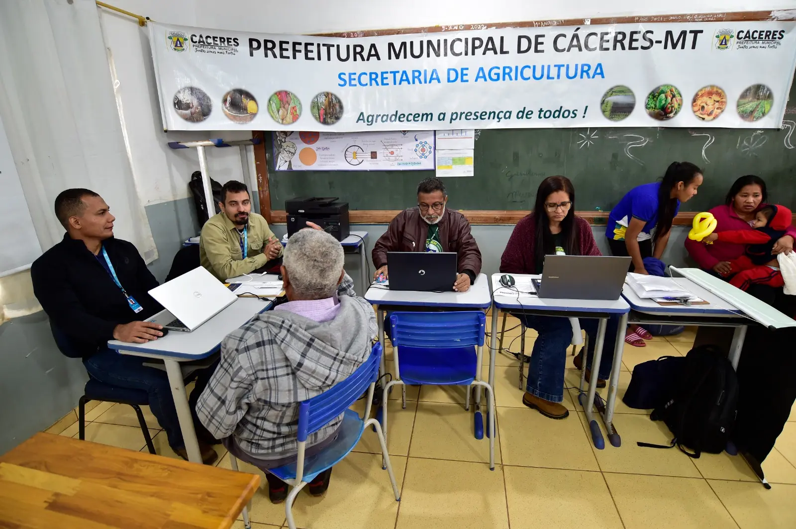 Imagem que mostra um sala de aula com mesas montadas com notebooks e servidores públicos atendendo pessoas. Na parede, há uma faixa com a divulgação da Secretaria de Agricultura de Cáceres.