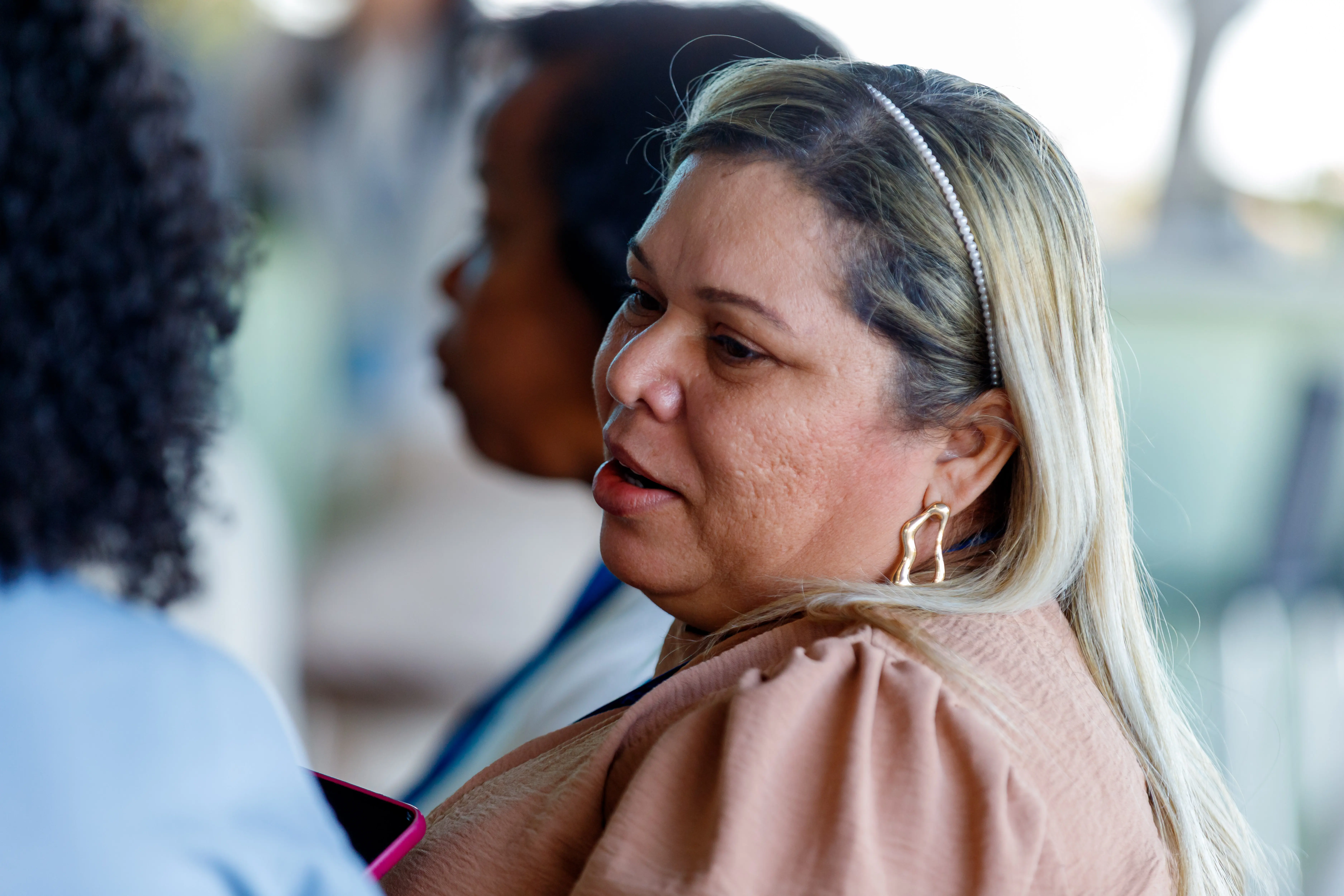 Mulher loira com tiara e blusa bege conversa atenta com outra pessoa. O foco está em seu rosto e expressão concentrada, em ambiente iluminado e informal