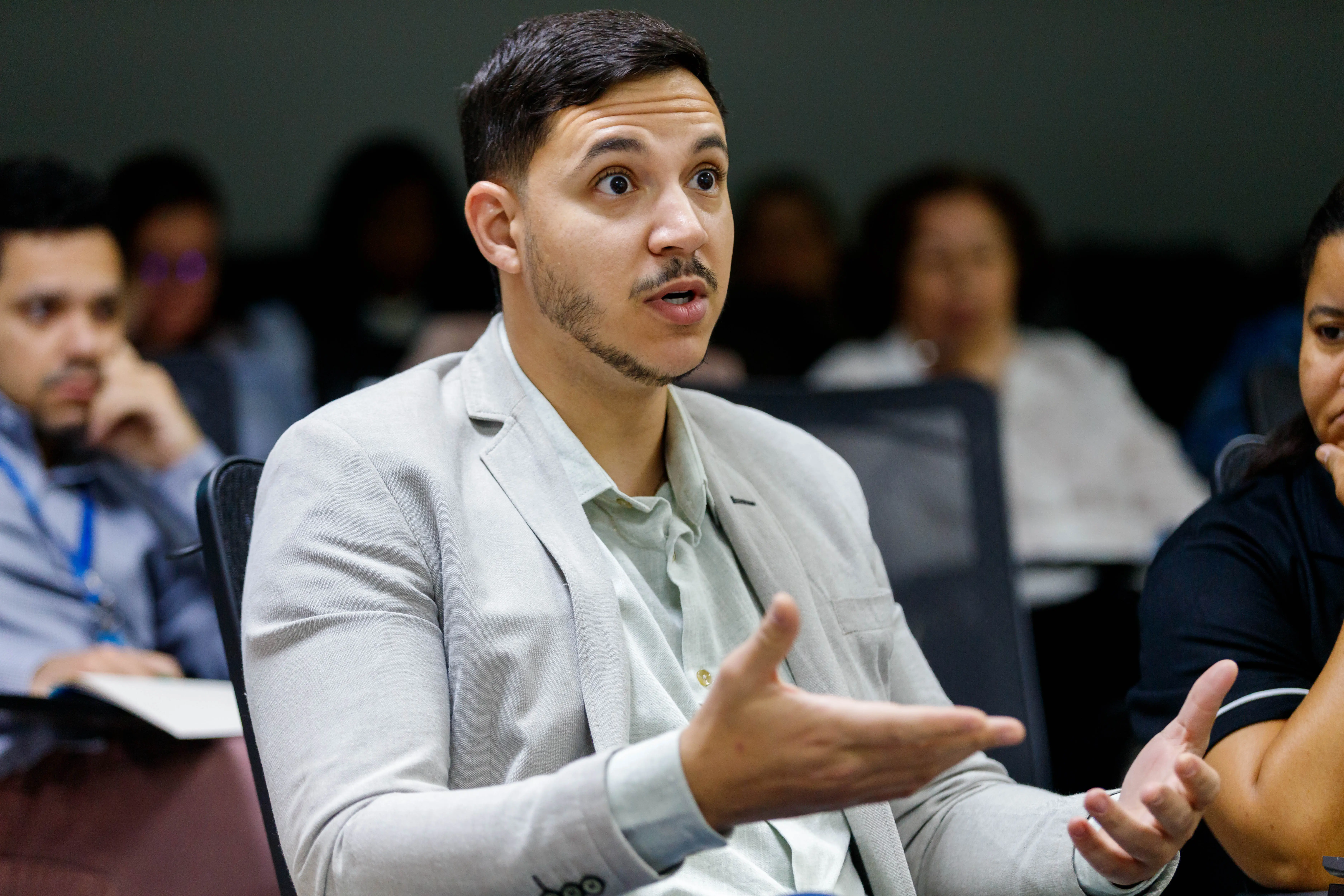 Homem de blazer claro e camisa verde fala com expressão de surpresa e gestos amplos, participando ativamente do debate em ambiente de capacitação