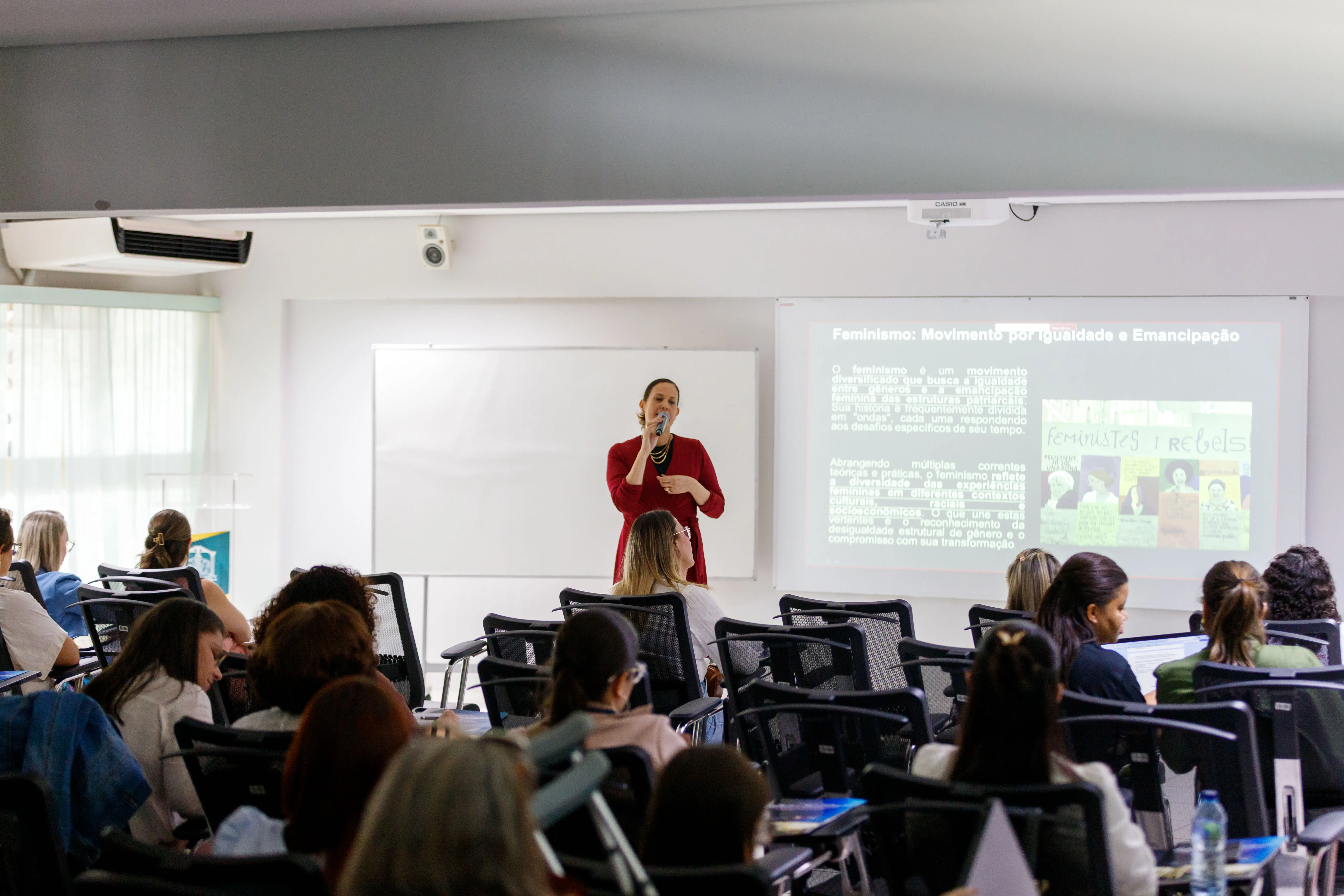 Mulher de vestido vermelho continua apresentação sobre igualdade de gênero, em sala cheia. A tela exibe o tema “Feminismo: Movimento por Igualdade e Emancipação”.