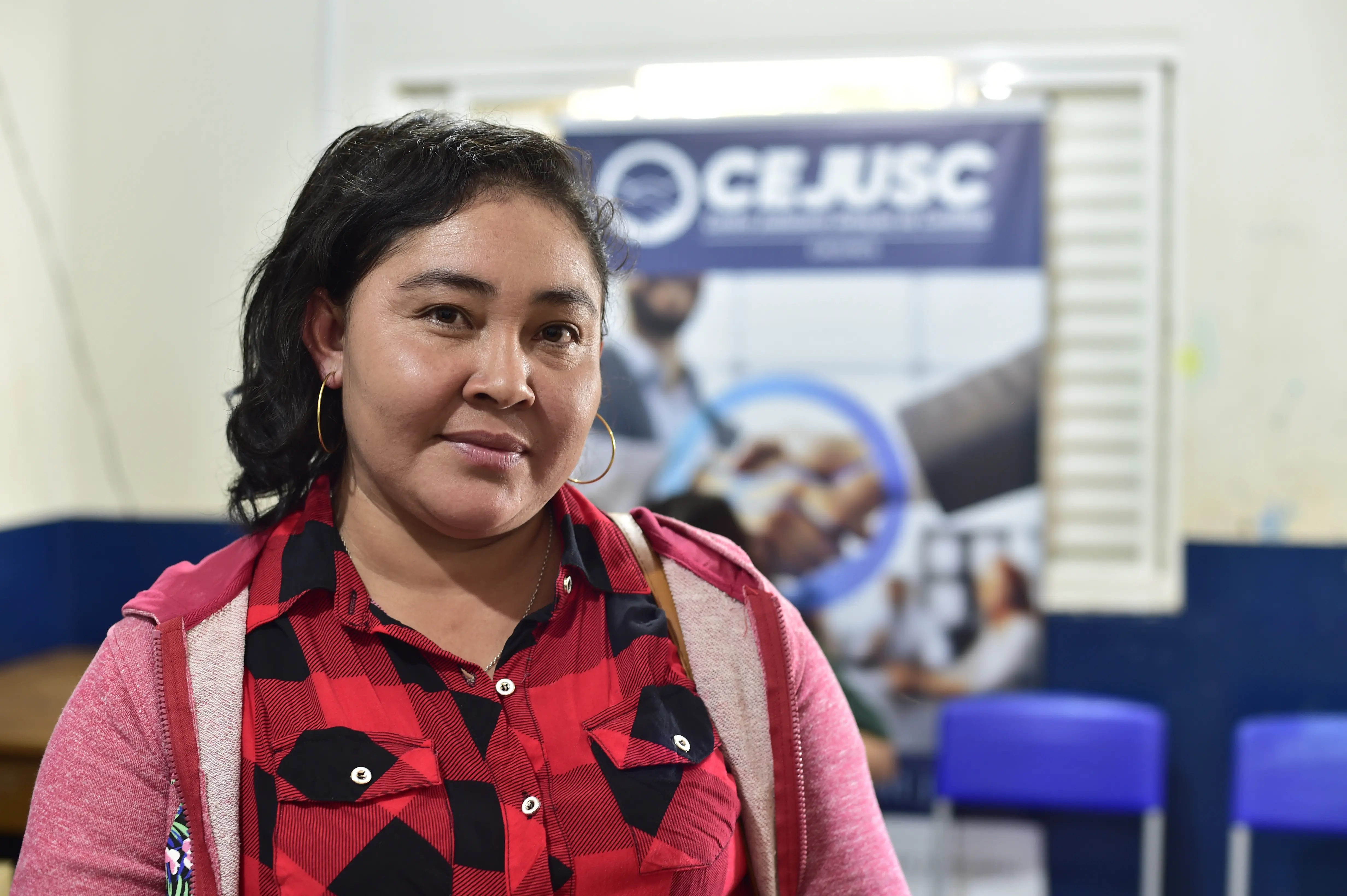 Mulher de expressão serena posa para a foto em frente a banner do Cejusc. Ela usa camisa vermelha xadrez e jaqueta rosa, em ambiente de mediação.