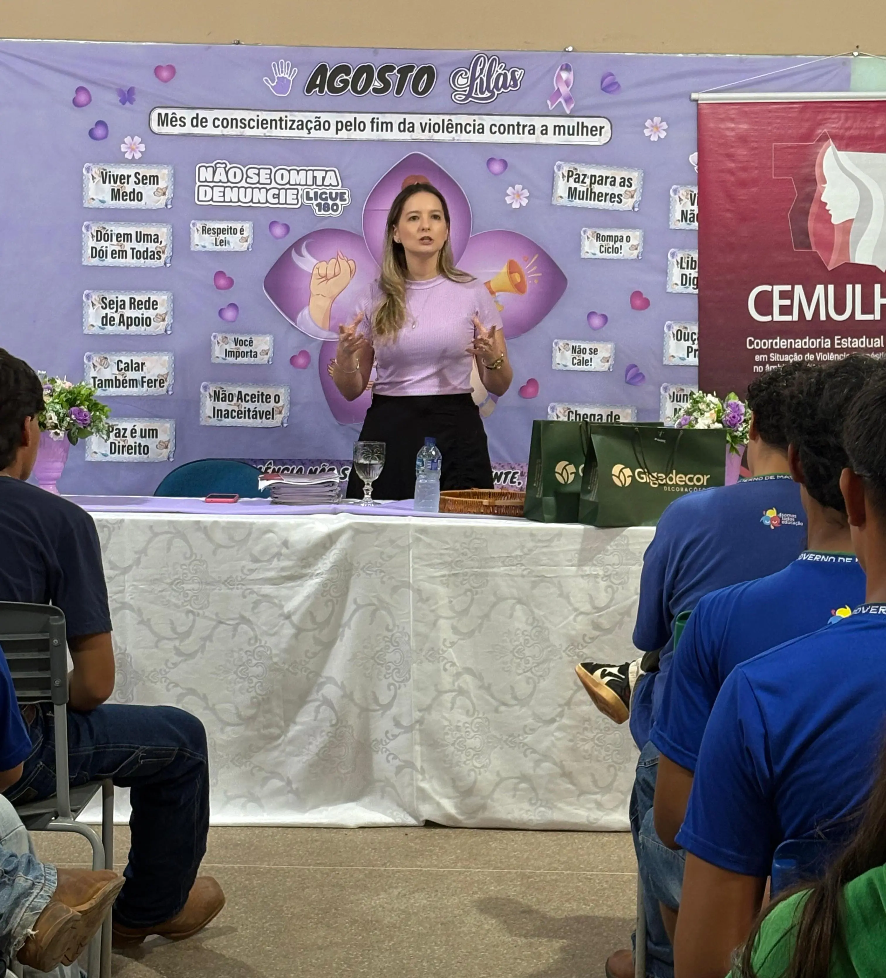 Juíza Djéssica Küntzer profere palestra a alunos de escola pública. Ela é uma mulher branca, loira, de olhos claros, usando blusa lilás. Atrás dela, há um painel na cor lilás com várias frases alusivas à campanha de combate à violência doméstica.