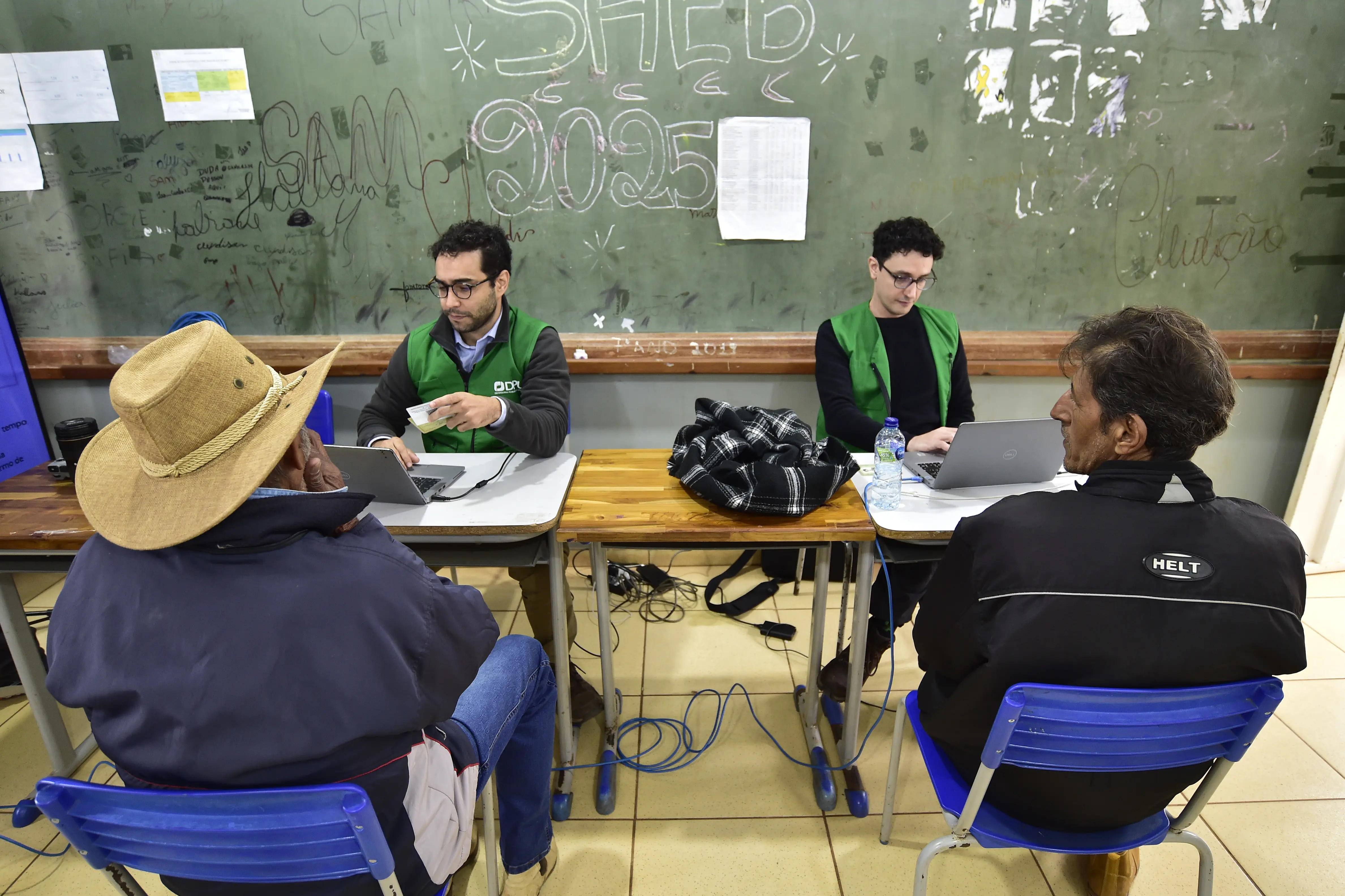 Foto que mostra dois defensores públicos da União, vestidos com jalecos verdes, atendendo, cada um, um homem, em uma sala de aula.