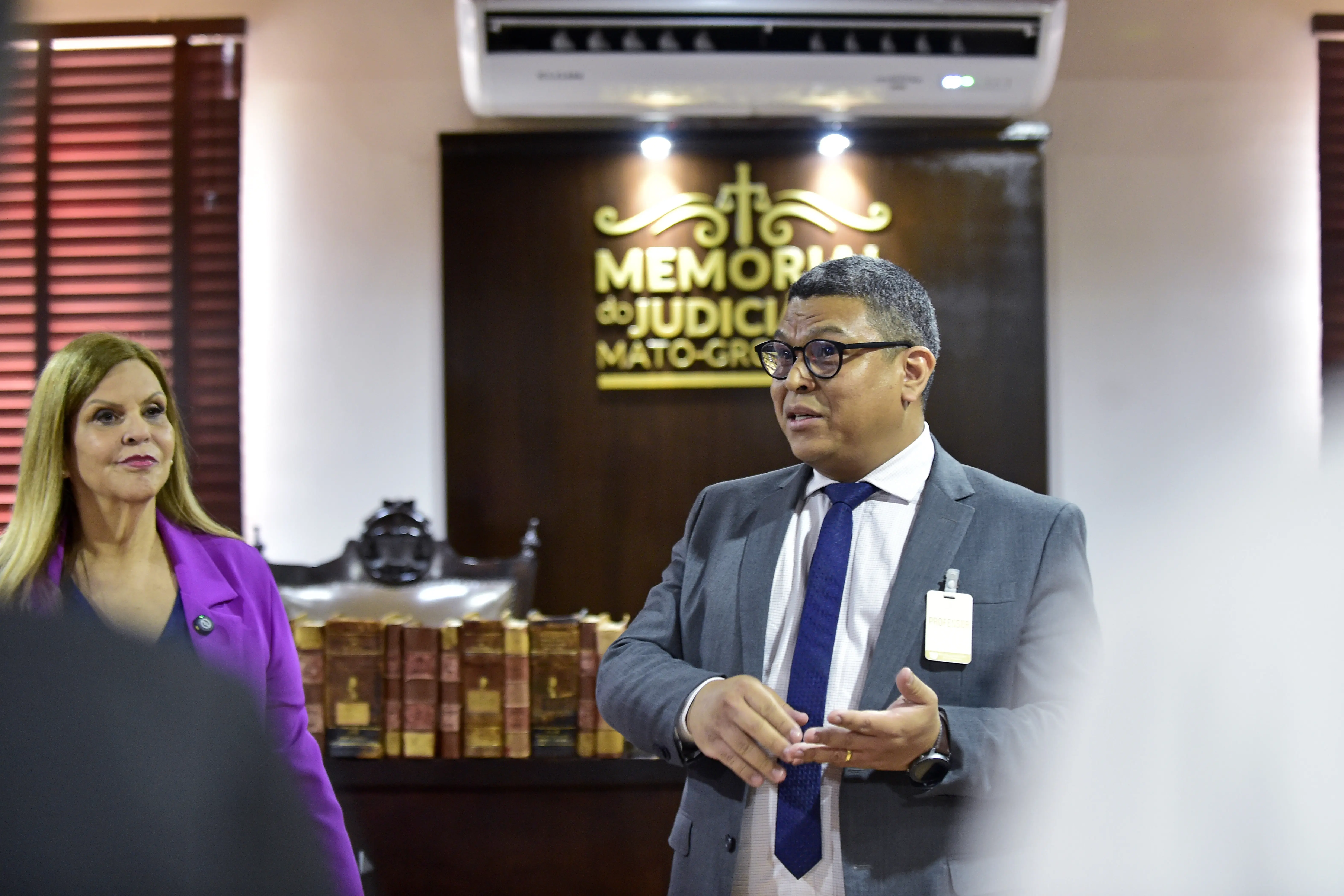 Homem de terno cinza fala em pé, gesticulando com as mãos, enquanto uma mulher de blazer roxo o observa ao lado. Ao fundo, livros antigos e o letreiro “Memorial do Judiciário Mato-grossense”.