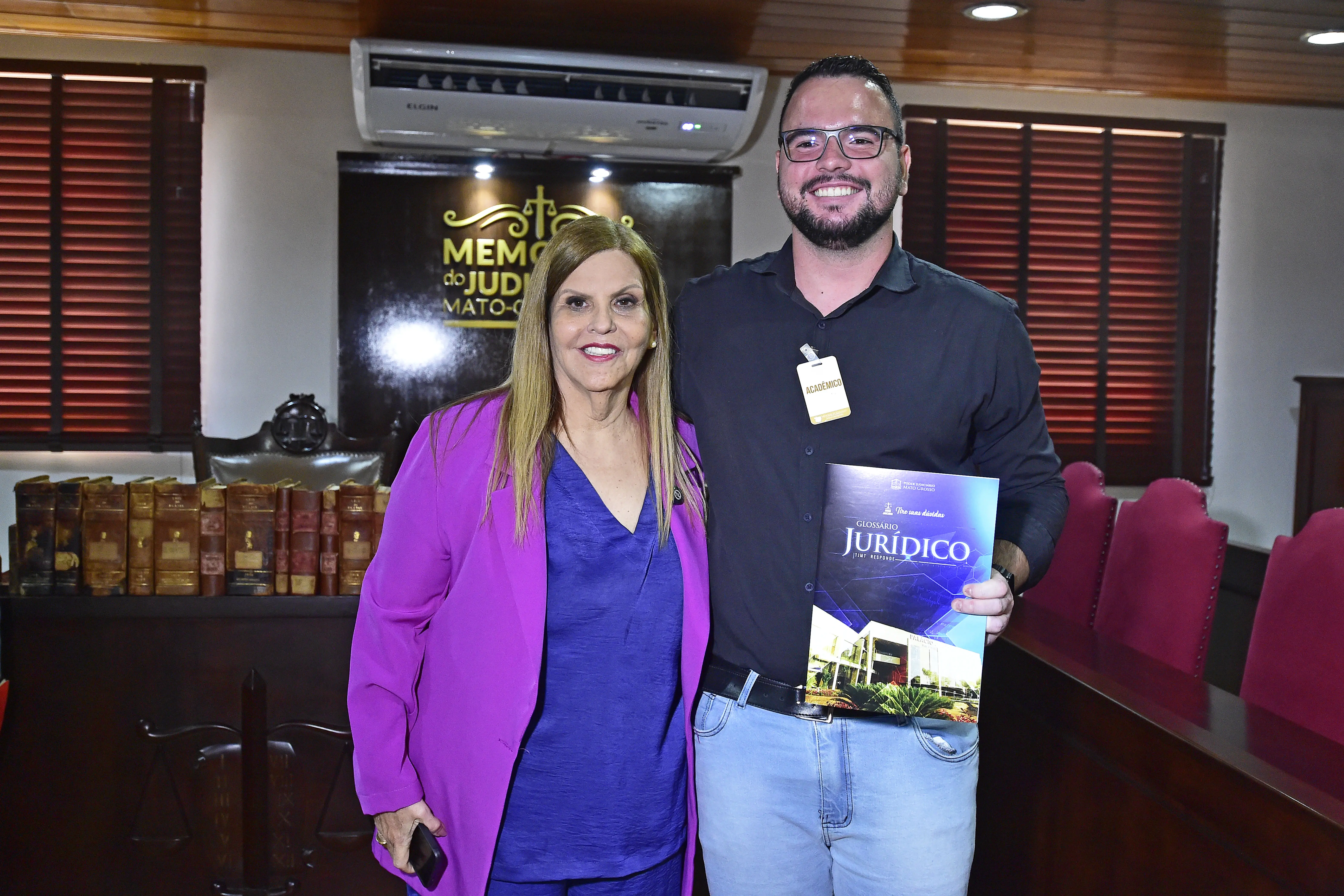 Mulher de blazer roxo e jovem de camisa preta posam juntos, sorrindo e exibindo exemplar do “Glossário Jurídico”. Atrás, o letreiro do Memorial e estantes de livros antigos completam o cenário.