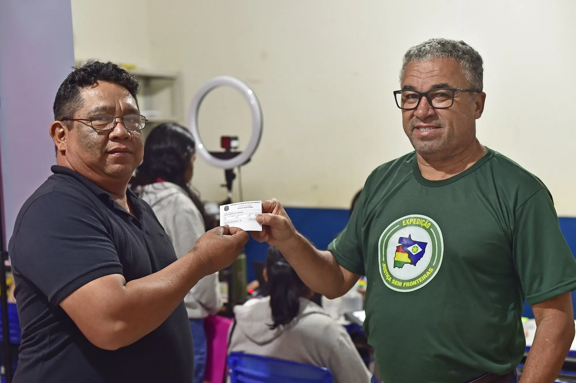 Foto mostra um cidadão recebendo seu título de eleitor das mãos do servidor do TRE, que está usando camiseta verde com logomarca da Justiça Sem Fronteiras. Eles sorriem para a foto.