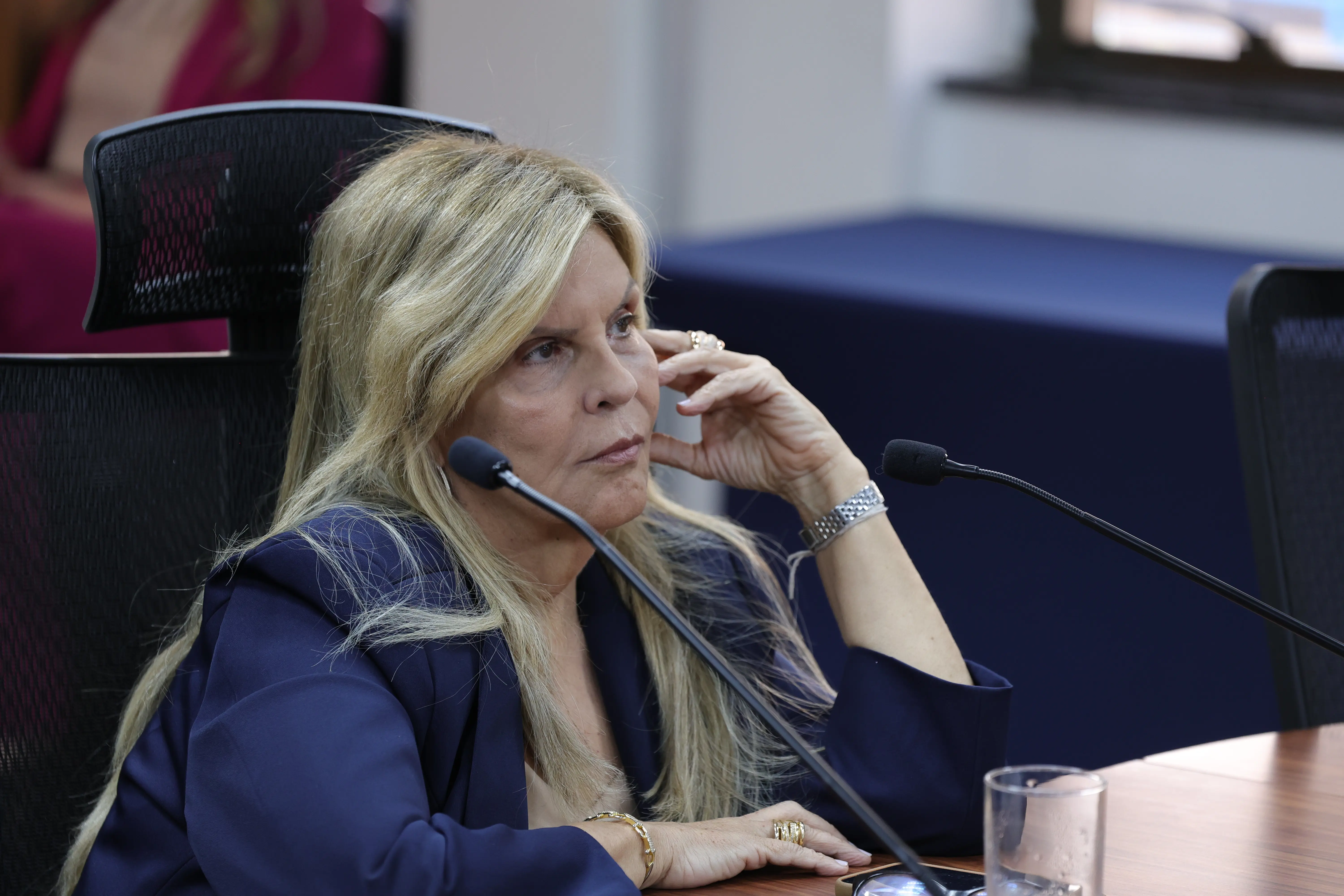Mulher loira, cabelos longos, veste blazer azul-marinho, toca o rosto com a mão direita enquanto observa ao lado, sentada entre dois microfones. Ao fundo, cadeira preta e mesa desfocada.