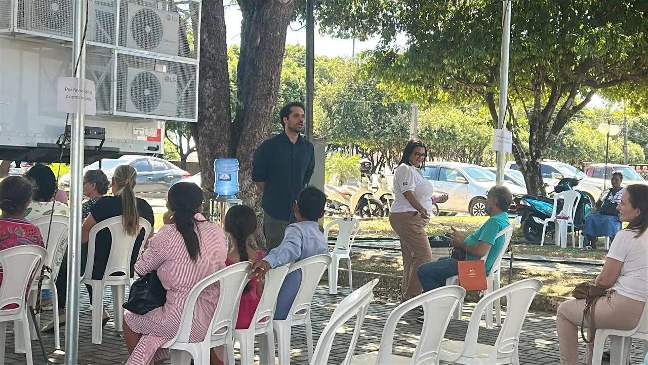 Foto horizontal em plano aberto que mostra diversas mulheres sentadas em cadeiras embaixo de uma tenda, ouvindo palestra do juiz Dante Aranha, que está à frente, num espaço aberto e arborizado da cidade de Paranaíta.