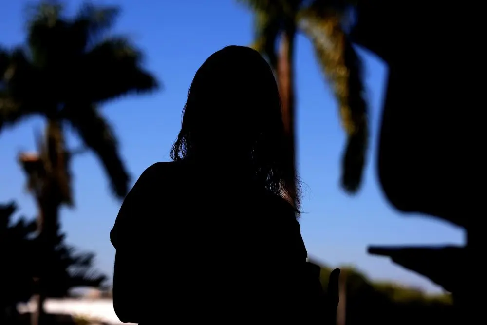 Foto horizontal que mostra a silhueta (sombra) de uma mulher vítima de violência doméstica. Ao fundo, aparece um céu azul e as sombras de coqueiros.