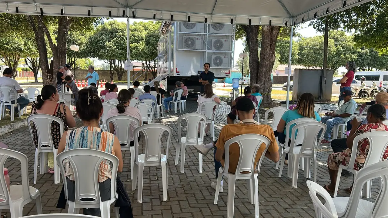 Foto horizontal em plano aberto que mostra diversas mulheres sentadas em cadeiras embaixo de uma tenda, ouvindo palestra do juiz Dante Aranha, que está à frente, num espaço aberto e arborizado da cidade de Paranaíta.