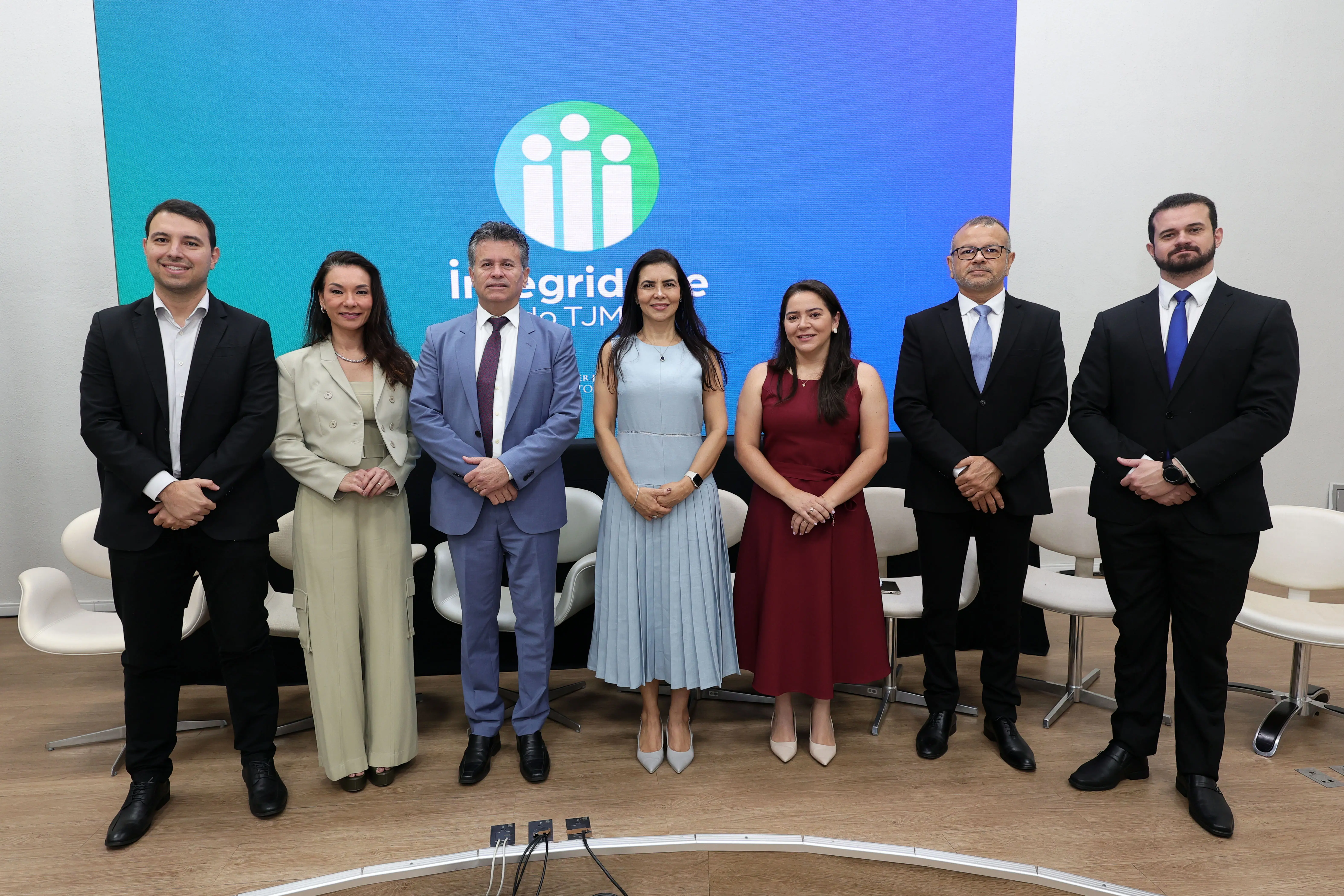 Sete pessoas estão em pé em uma fileira no palco, posando para uma foto oficial. Elas estão vestidas formalmente em ternos e vestidos, em frente a um painel de LED azul com o logo "integridade do TJMT". Cadeiras brancas estão atrás delas.