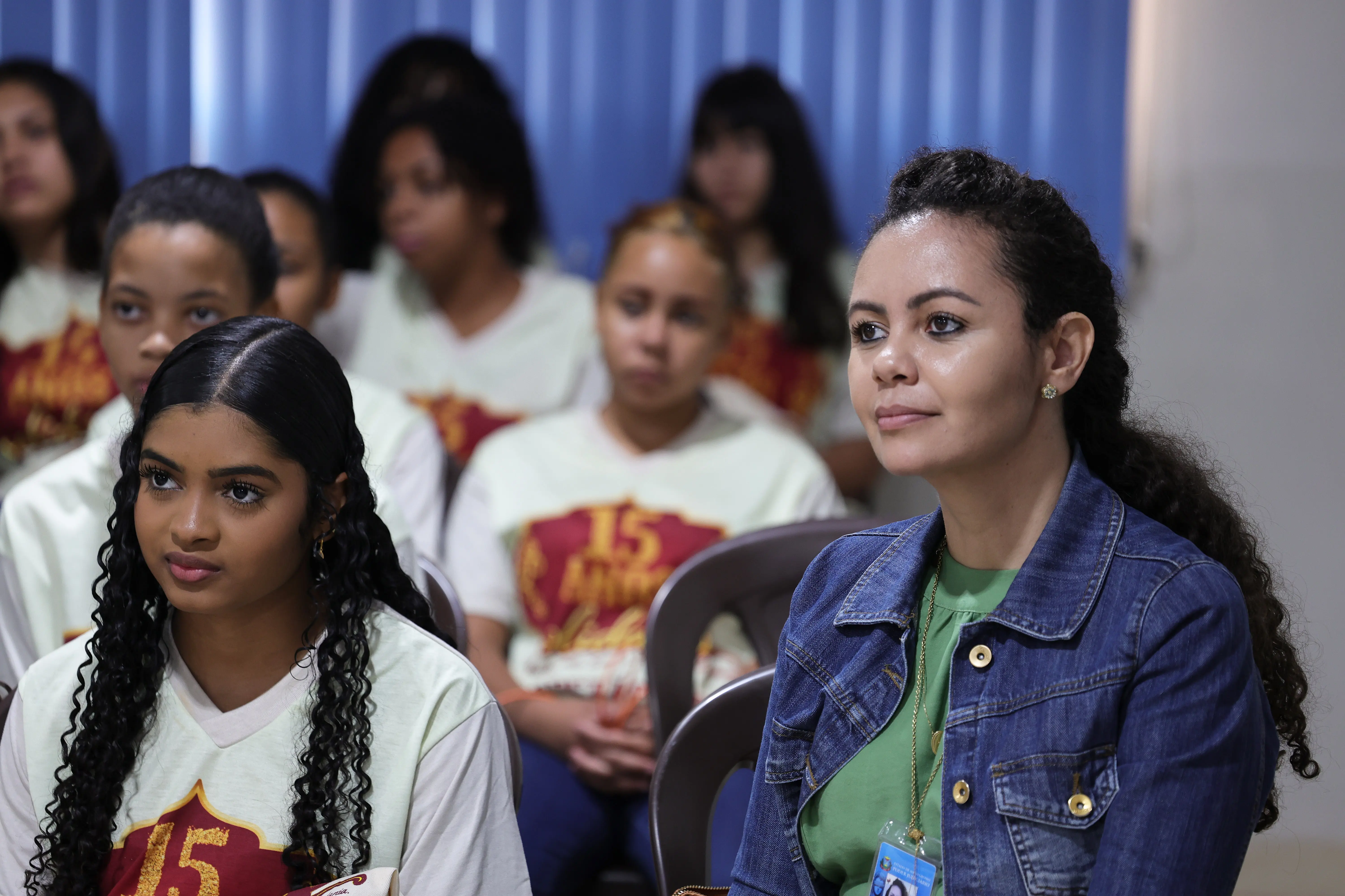 Mulher de jaqueta jeans atenta em primeiro plano. Ao lado, jovem de cabelos cacheados compridos e, ao fundo, outras pessoas sentadas na plateia assistem ao evento.