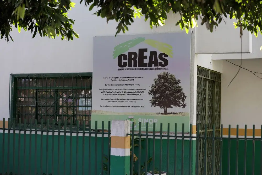 Foto horizontal que mostra a fachada do CREAS de Sinop. É uma casa com parede pintada de branco verde e listra amarela, com a placa do Centro de Referência Especializado de Assistência Social. Na parte superior aparecem folhas de árvores.