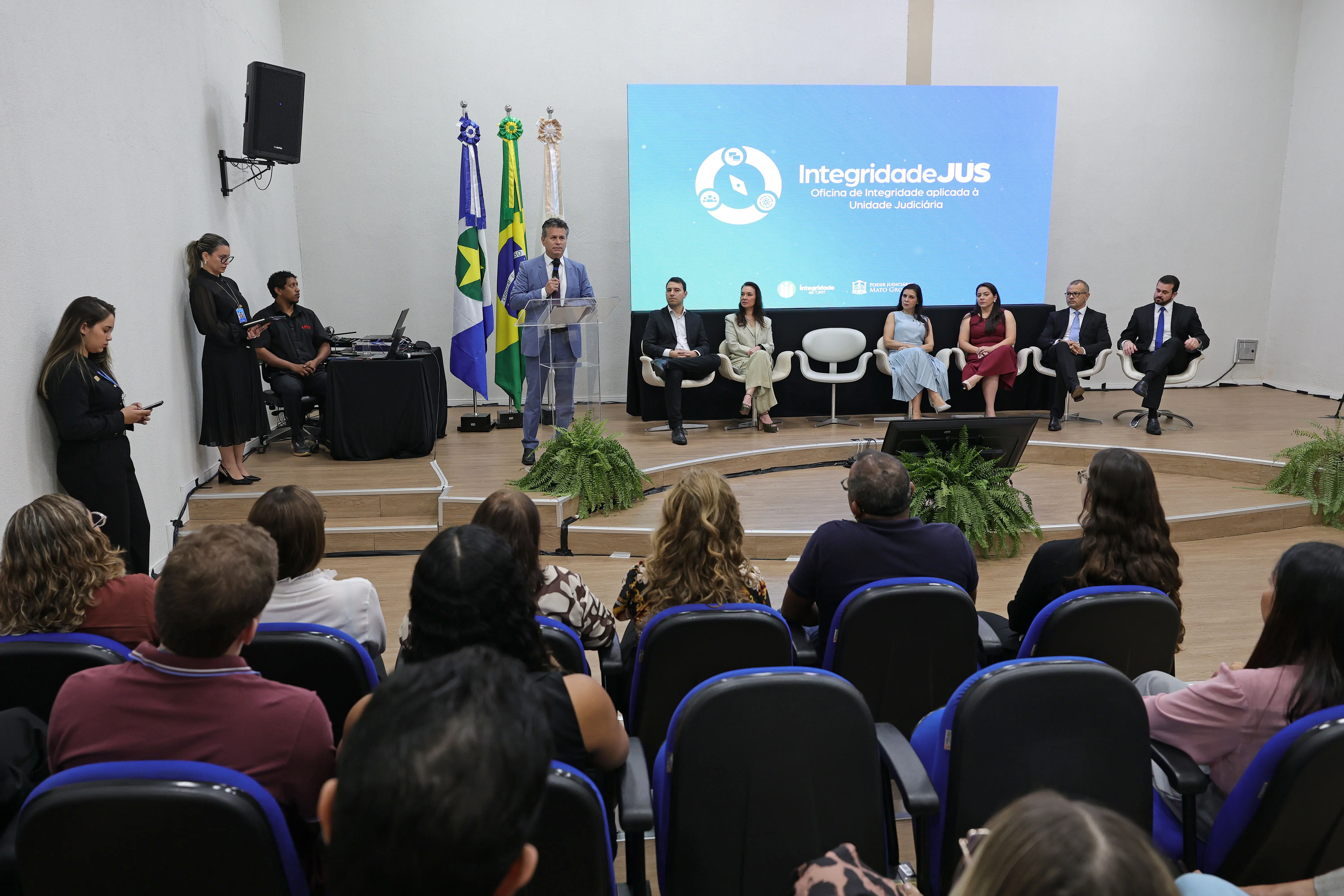 Em um auditório, várias pessoas estão sentadas em cadeiras azuis em fileiras escalonadas. Elas assistem a uma apresentação, e em primeiro plano, uma tela exibe a logo "integridade do TJMT". A plateia é diversificada e veste roupas formais e casuais.