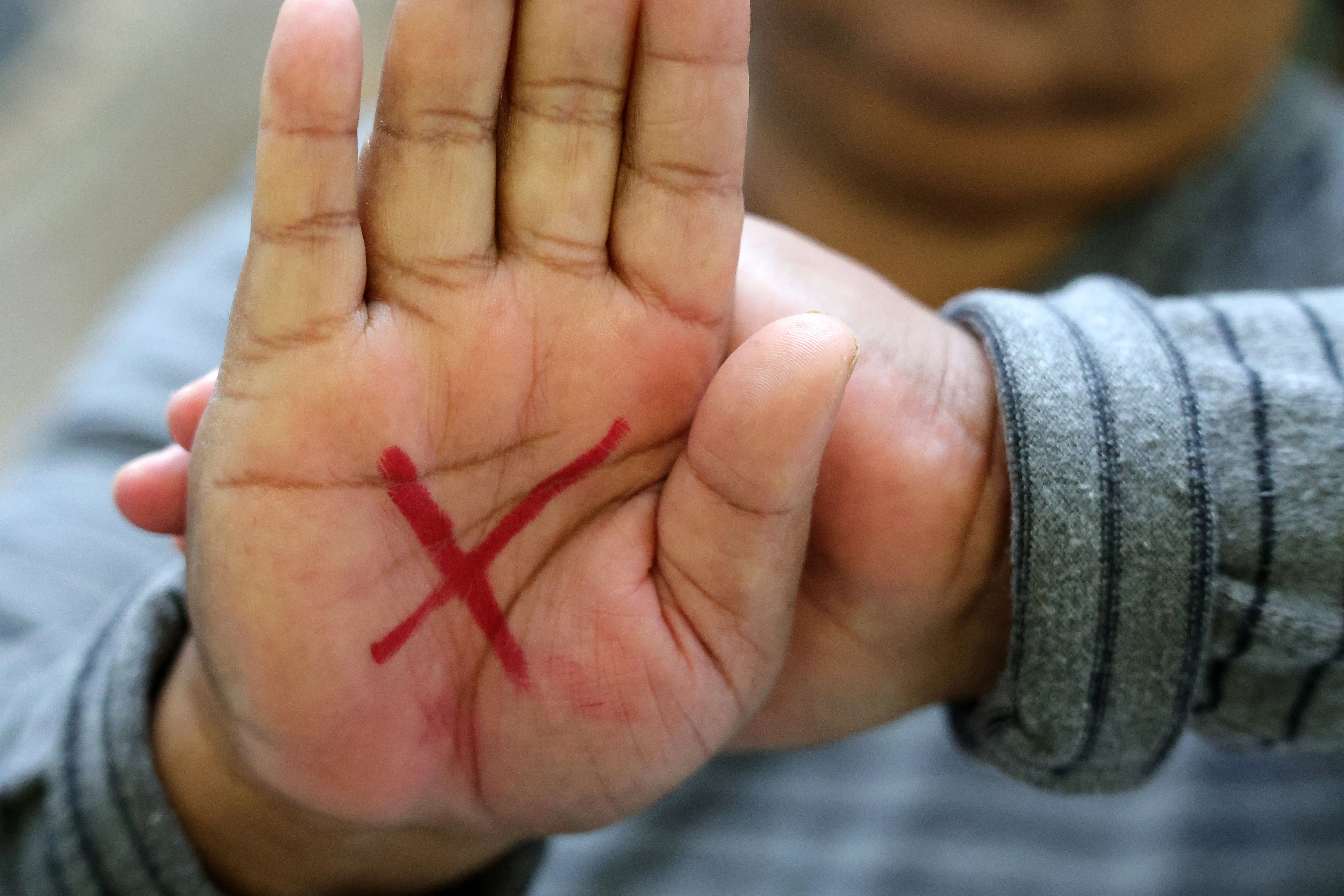 Close de uma mão com um X vermelho desenhado na palma, símbolo de combate à violência contra a mulher. Ao fundo, outra mão e manga de blusa cinza