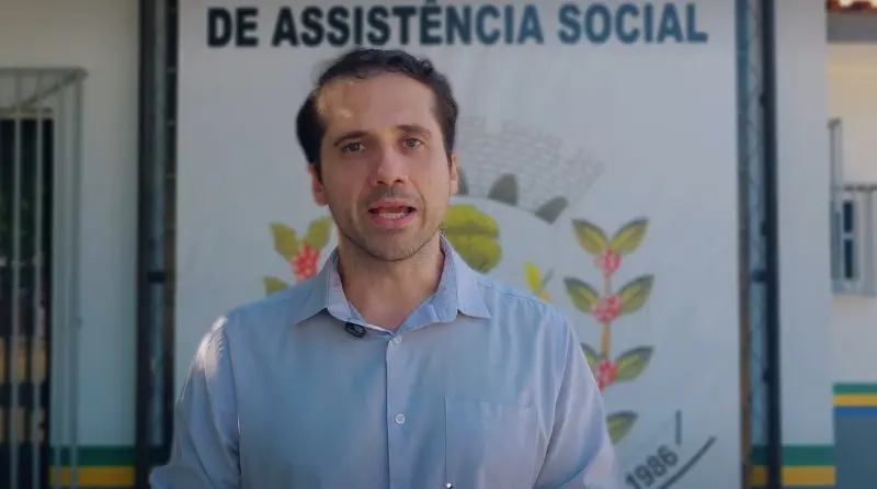 Print de tela de um vídeo que mostra o juiz da comarca de Paranaíta, Dante Rodrigo Aranha, falando em direção à câmera. Ele é um homem jovem, branco, de cabelo castanho claro, olhos claros, usando camisa social azul clara. Ele está em frente à fachada da Secretaria de Assistência Social. 