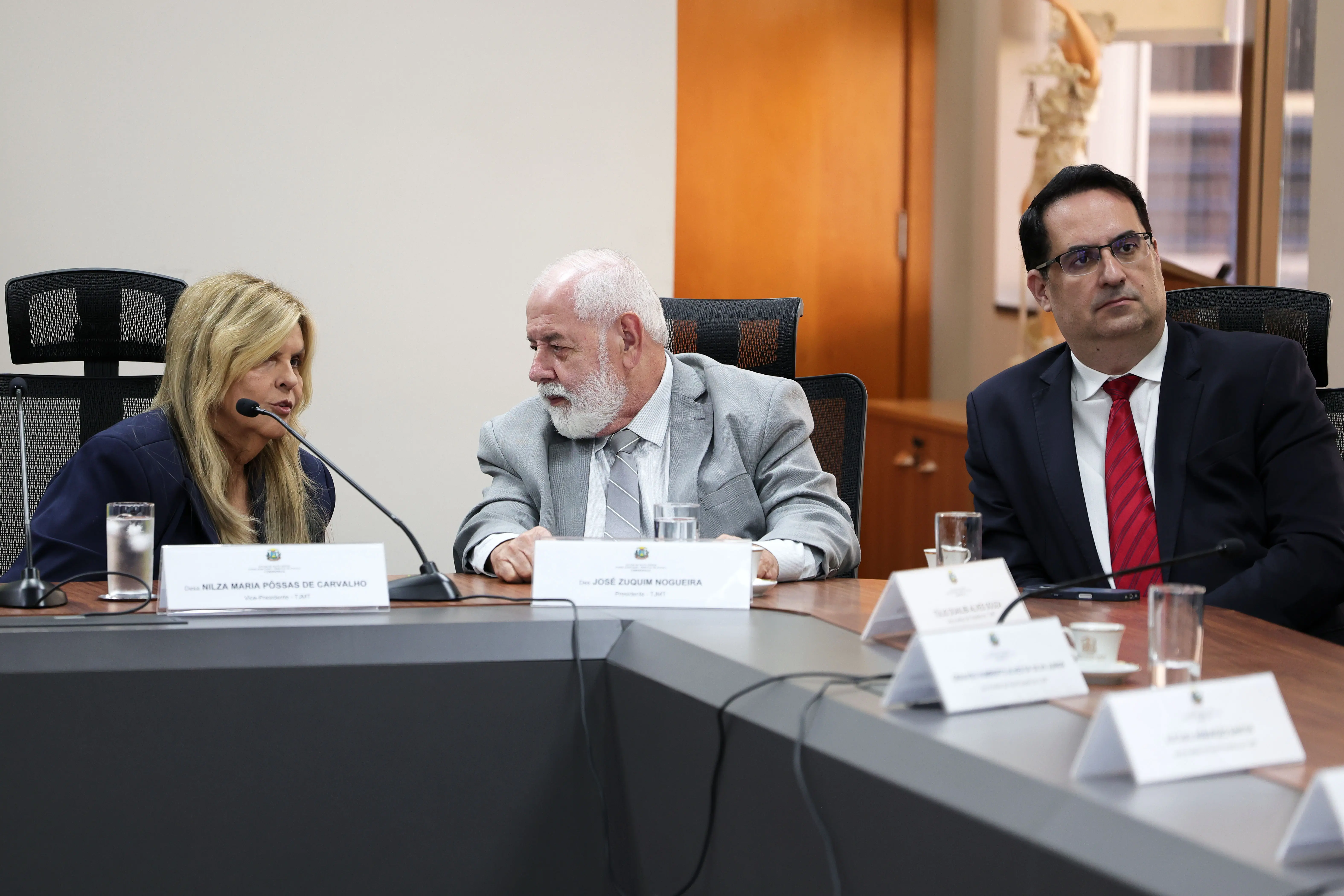 Três pessoas sentadas à mesa: mulher loira fala ao microfone, ao centro homem de barba branca e terno cinza, à direita homem de óculos, terno escuro e gravata vermelha. Ao fundo, estátua da Justiça.