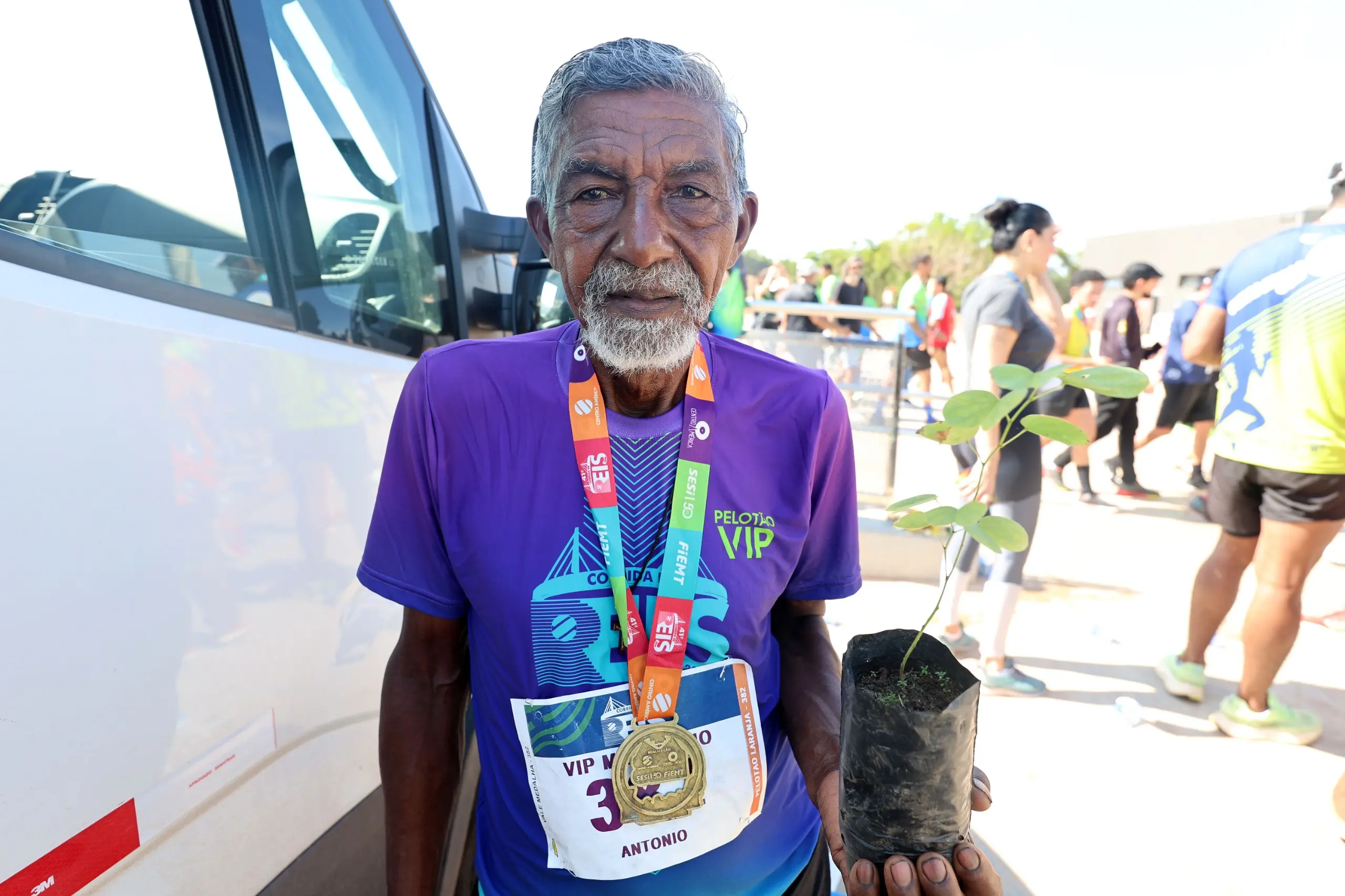 Homem idoso, de camisa roxa do evento e medalha no peito, posa em área externa da corrida, segurando uma muda de árvore em saco plástico. Ao fundo, veículo de apoio e participantes circulam após a prova.