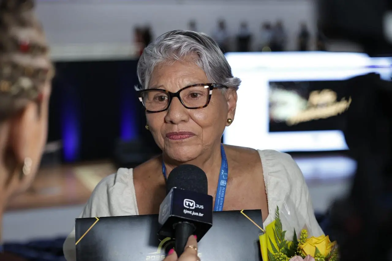 Mulher idosa de cabelos brancos e óculos, com um buquê de flores e um certificado escuro. Ela está sendo entrevistada por um microfone da 