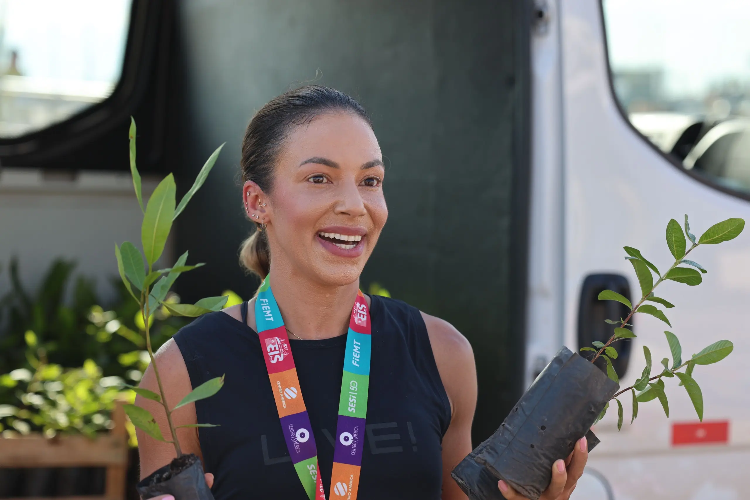 Mulher com camiseta preta e medalha no pescoço segura duas mudas de árvores, uma em cada mão. Ela conversa animadamente em frente a uma van, com caixas de plantas visíveis ao fundo.