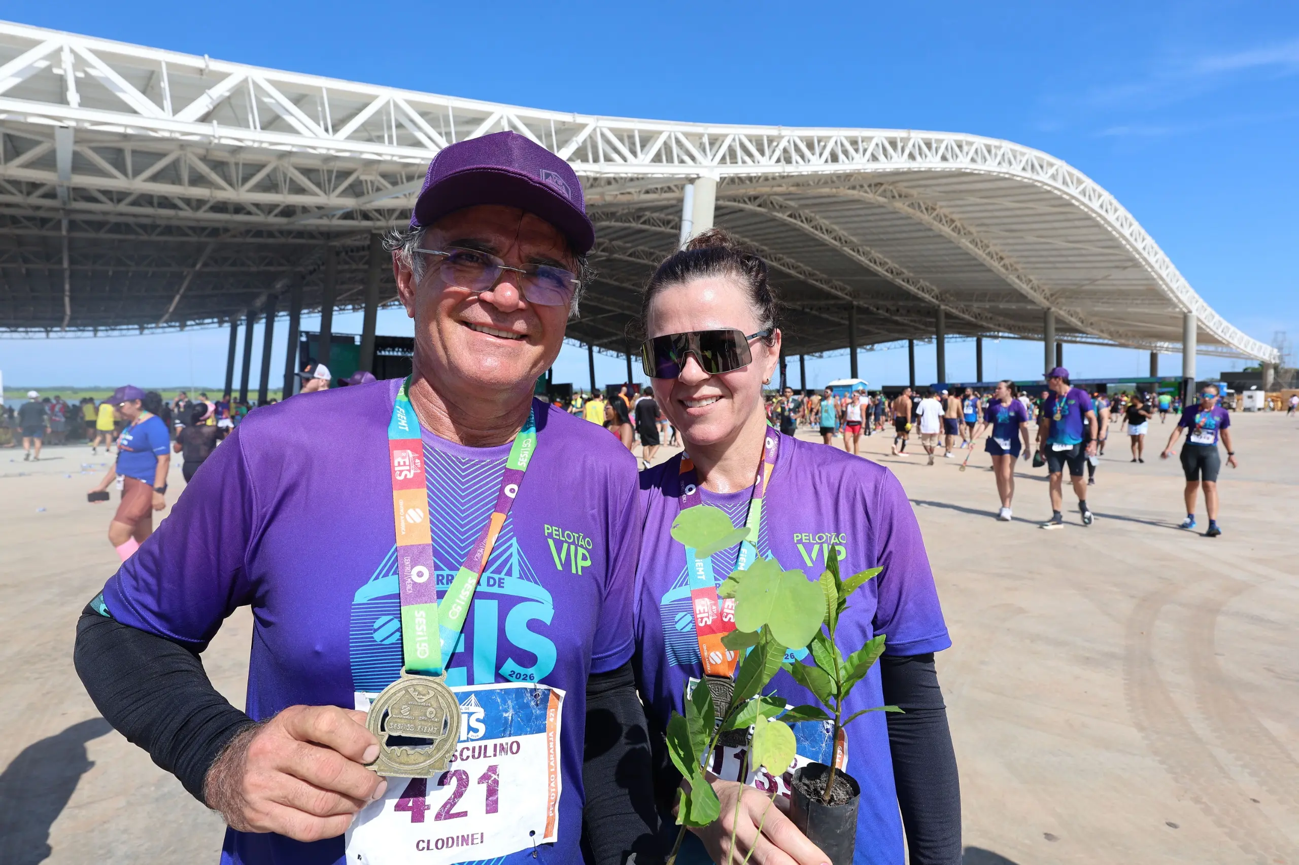 Casal de corredores usa camisetas roxas do evento e medalhas, sorrindo para a foto enquanto segura uma muda de árvore. Ao fundo, pavilhão coberto e público disperso na área da corrida.