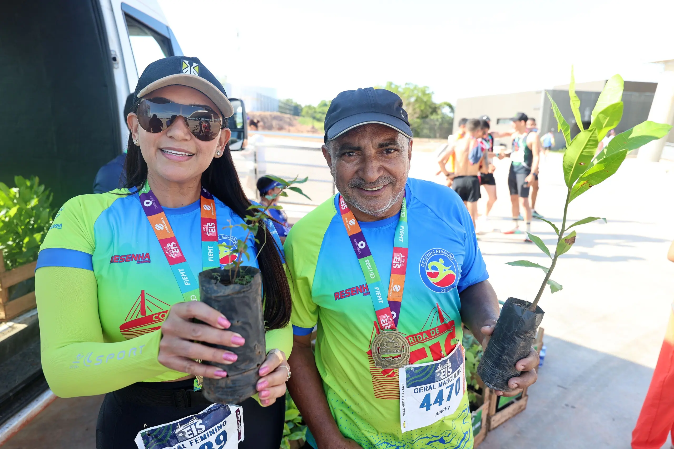Mulher e homem posam lado a lado após a corrida, usando camisetas verdes do grupo esportivo, medalhas no pescoço e segurando mudas de árvores. A cena ocorre ao lado de uma van de apoio, em área externa do evento.