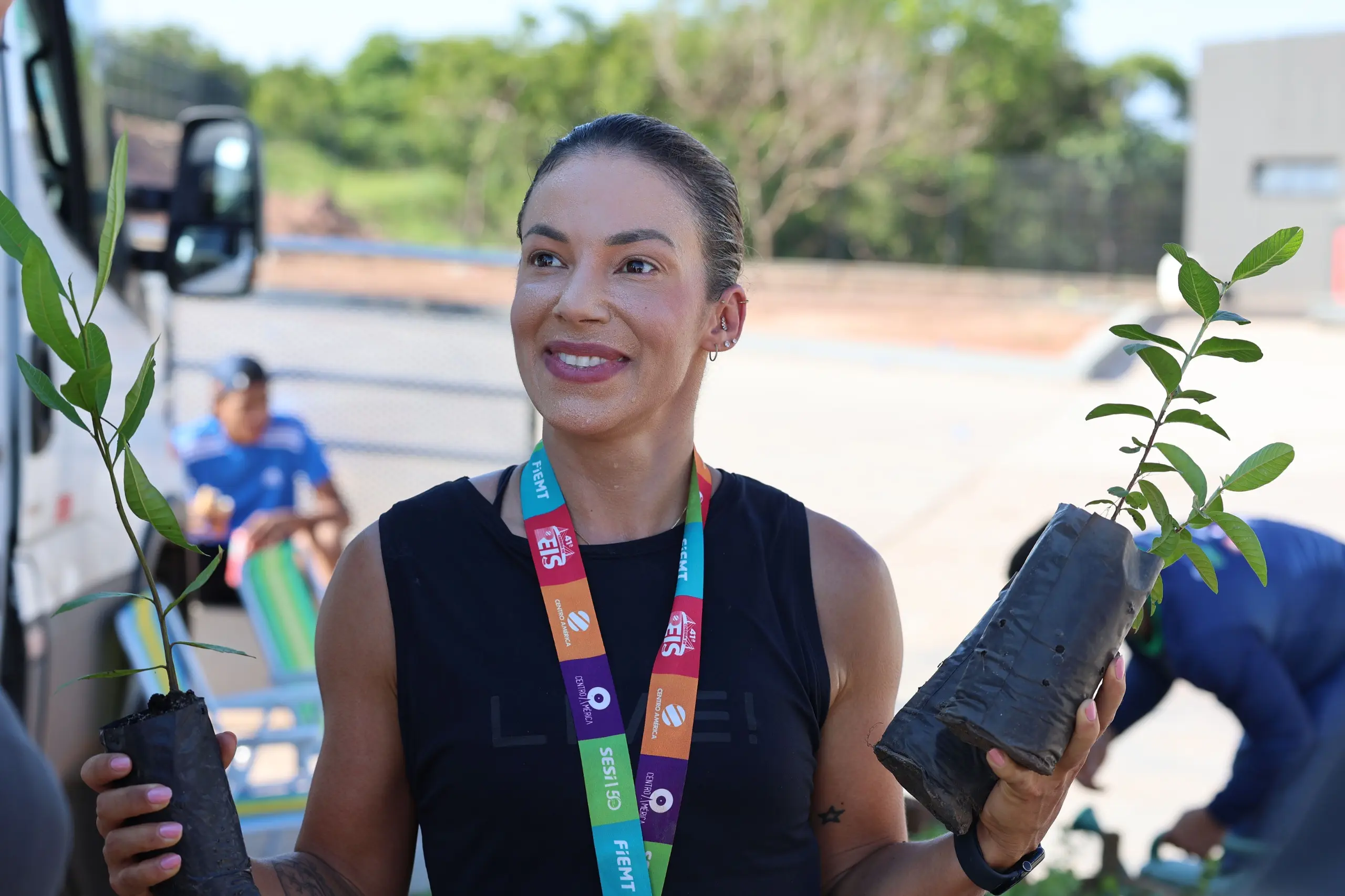 Mulher posa sorrindo com duas mudas de árvores nas mãos, usando medalha e roupa esportiva. A imagem foi registrada em espaço externo, próximo à área de retirada das plantas.