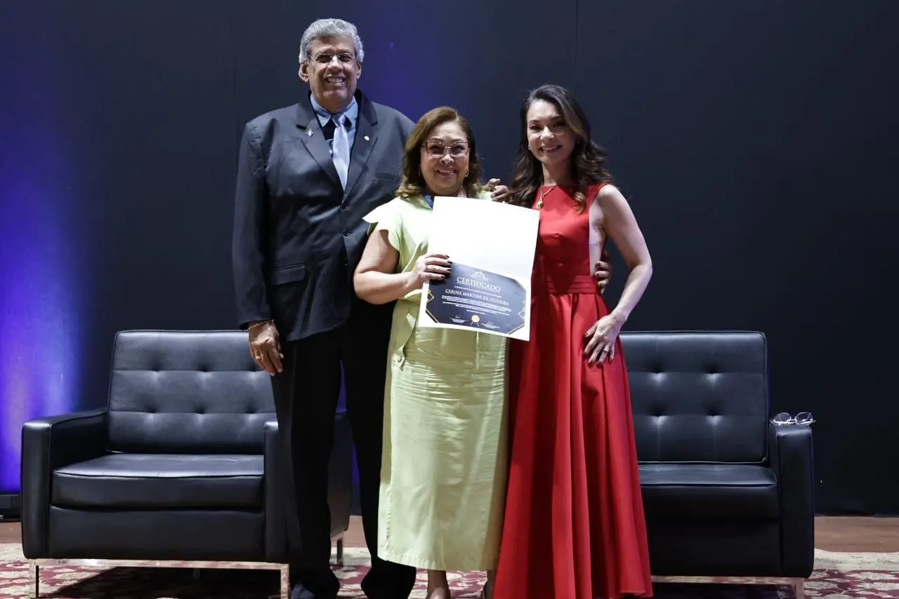 Três pessoas em um palco: um homem de terno cinza, uma mulher de vestido verde-claro segurando um certificado e uma mulher de vestido vermelho. Estão em frente a um sofá de couro preto e fundo escuro.