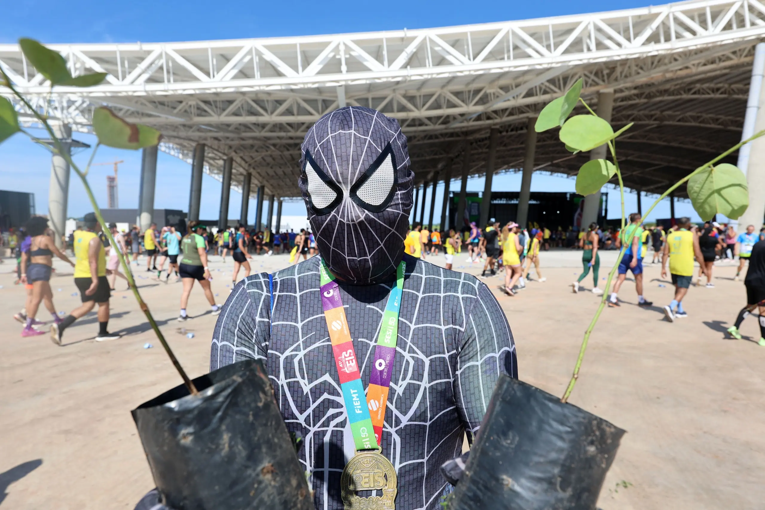 Participante fantasiado de super-herói, com medalha no pescoço, segura duas mudas de árvores. A cena ocorre em amplo espaço aberto, com estrutura metálica ao fundo e diversos corredores caminhando.