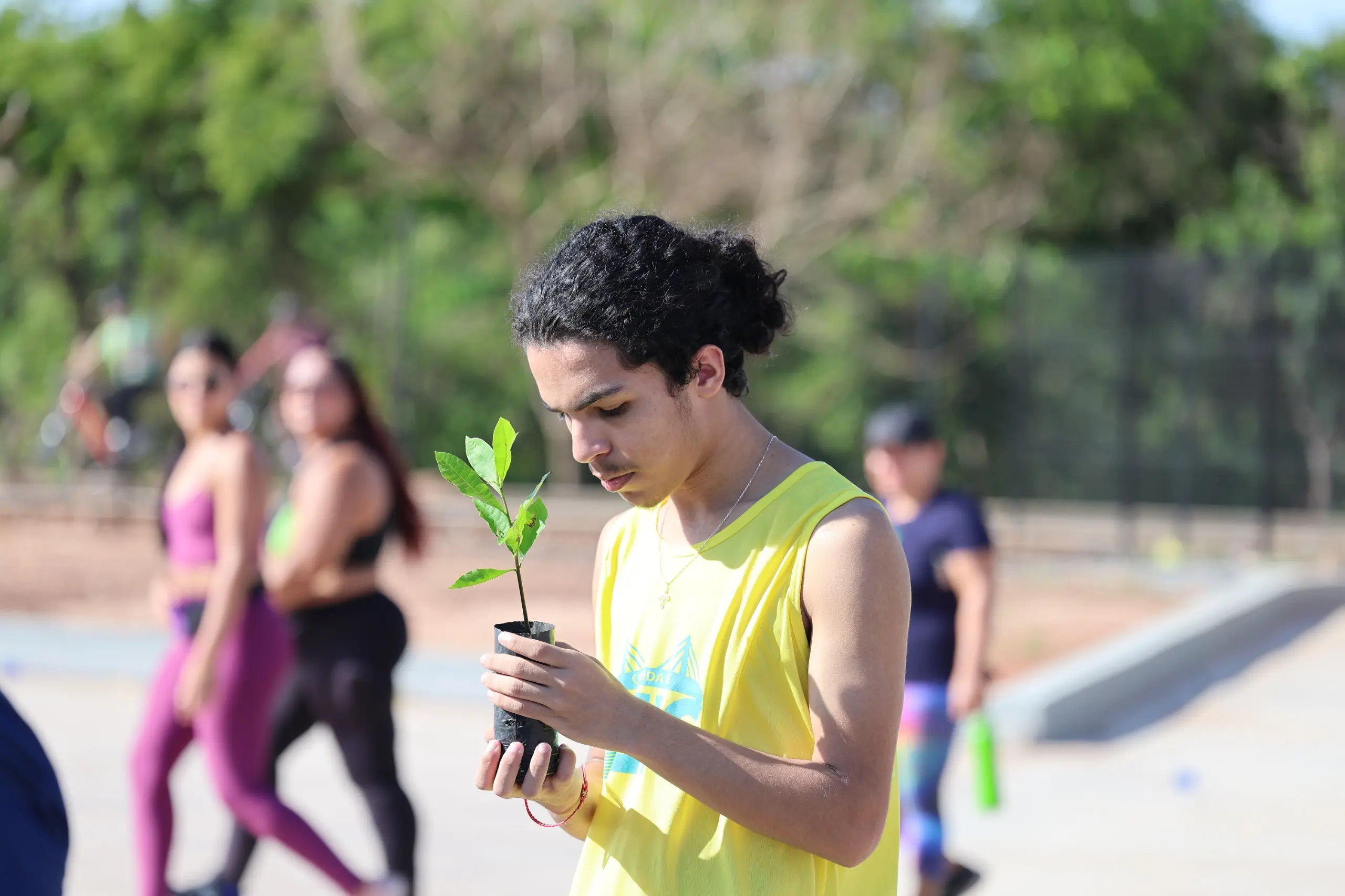 Jovem participante observa atentamente uma muda de árvore que segura nas mãos. Ao fundo, outros corredores caminham pela área aberta do evento, com vegetação e pista visíveis.