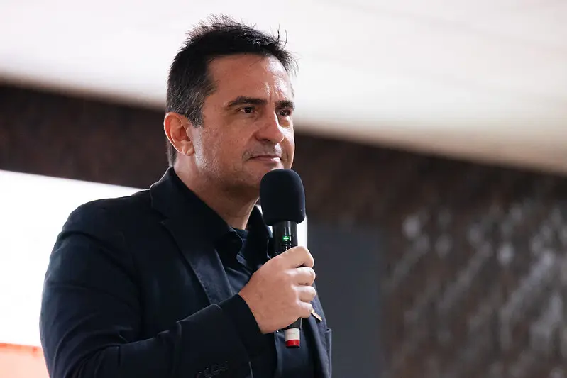 Foto horizontal que mostra o promotor de justiça Luciano Furtado Loubet do busto pra cima, falando ao microfone, no palco do auditório do TJMT. Ele é um homem branco, de cabelo e olhos escuros, usando camisa e blazer pretos. 