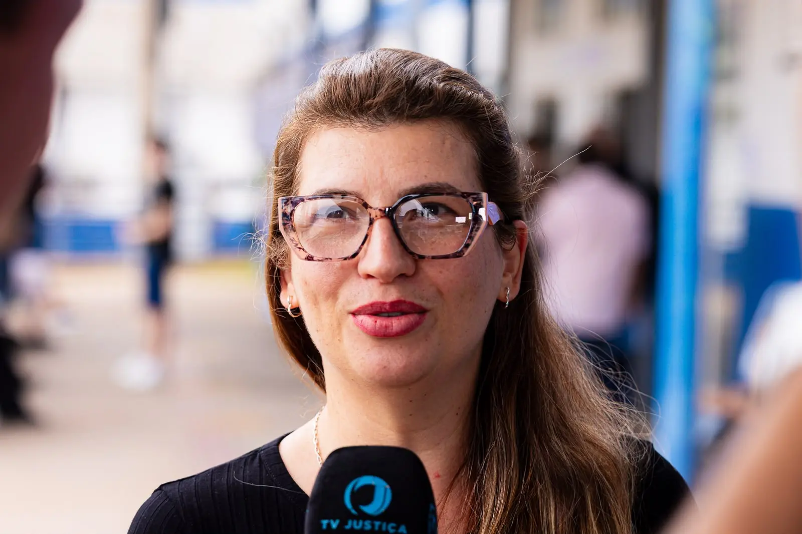 Mulher de cabelos castanhos e óculos de grau sorri de perfil para a esquerda enquanto fala próximo a um microfone da "TV JUSTIÇA". O fundo mostra árvores e estruturas ao ar livre fora de foco.