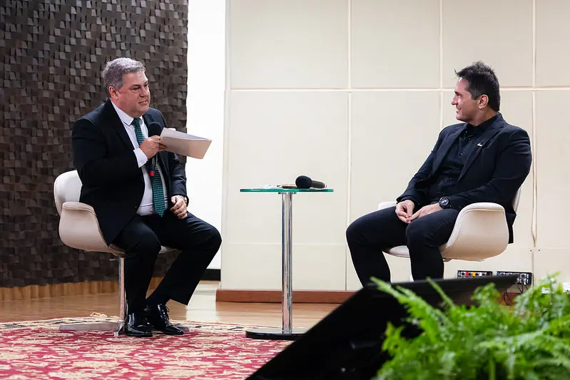 Foto horizontal em plano aberto que mostra o desembargador Wesley Sanchez e o promotor de justiça Luciano Loubet sentados em poltronas off-white, no palco do auditório do TJMT. O primeiro fala ao microfone e olha para o interlocutor. No canto inferior direito, aparece uma samambaia compondo o palco.