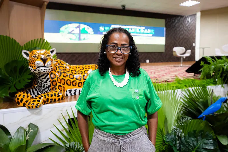 Foto horizontal que mostra a presidente da Asmats e educadora ambiental Cidinha Nascimento posando para a foto em frente ao palco do auditório do TJMT. Ela é uma mulher negra, de cabelo preto, cacheado, na altura do ombro, usando camiseta verde da Asmats, calça cinza, colar branco e óculos de grau.